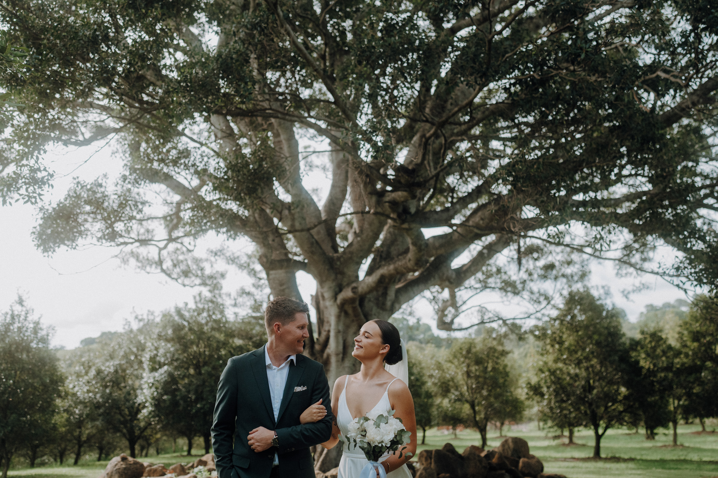 A couple in formal attire stands together, smiling under a large tree in a scenic outdoor setting. The woman holds a bouquet.