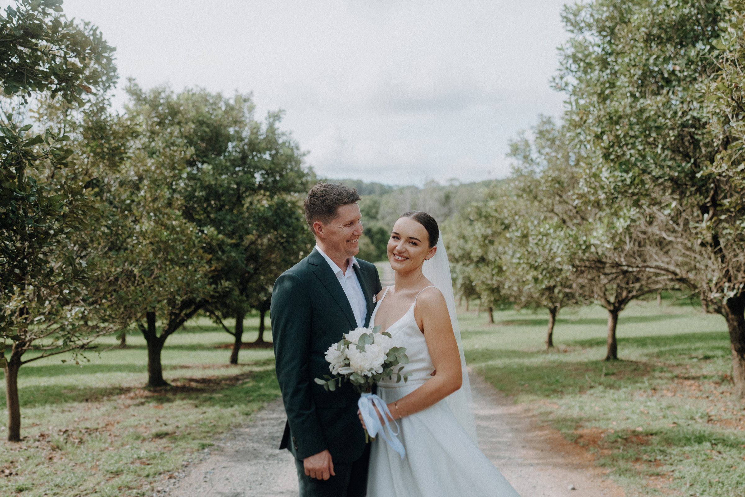 A couple poses outdoors on a tree-lined path, with the woman in a white wedding dress holding a bouquet and the man in a dark suit.