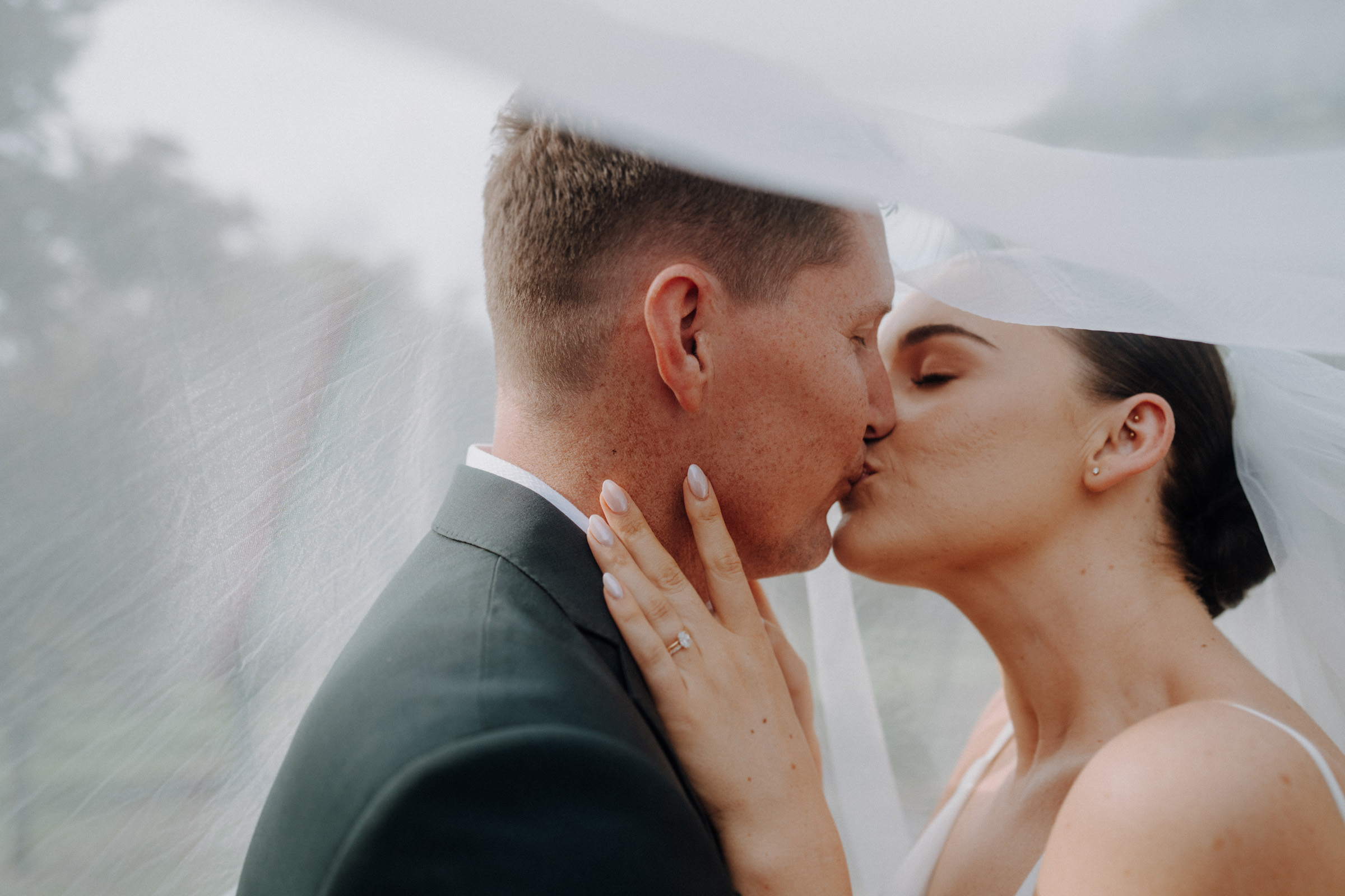 A bride and groom share a kiss under a veil outdoors. The bride's hand is on the groom's neck, showing her engagement ring.