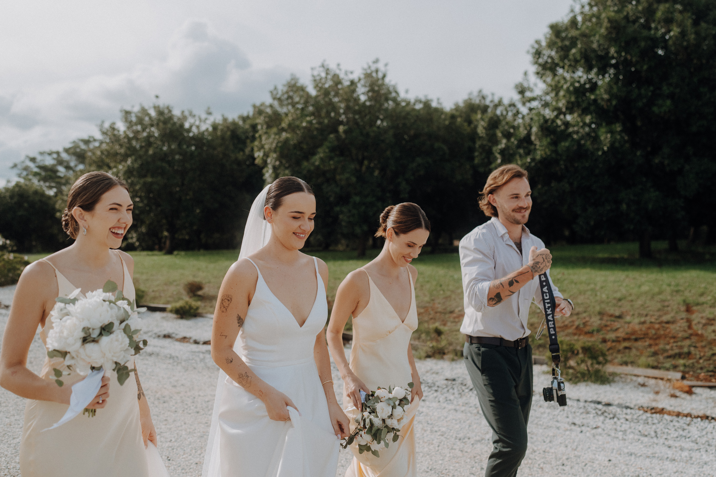 A groom and three bridesmaids walk outdoors on a gravel path, holding bouquets. The groom carries a camera strap. Trees and a cloudy sky are in the background.