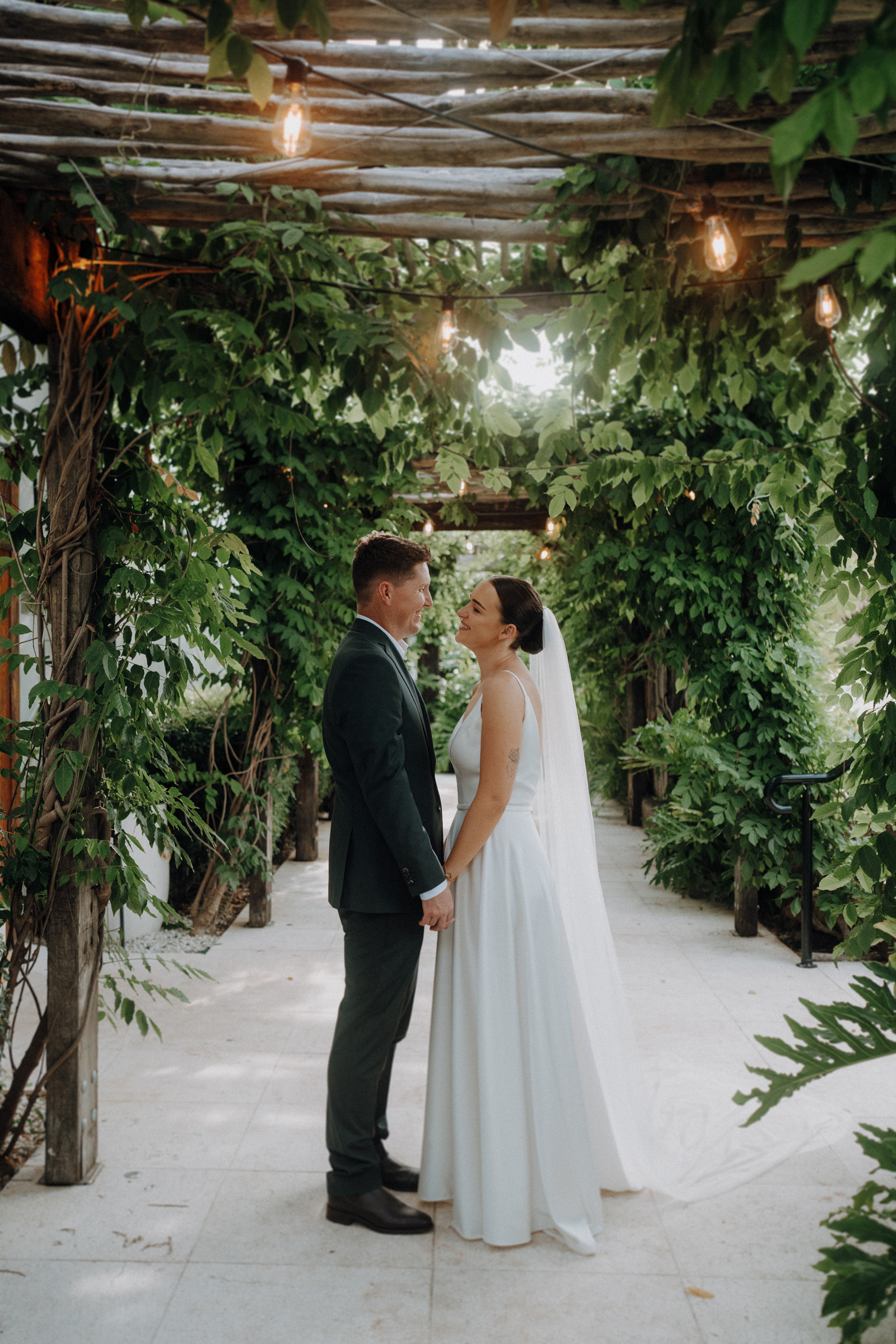 A bride and groom stand under a leafy pergola, facing each other and holding hands, with soft lighting above them.