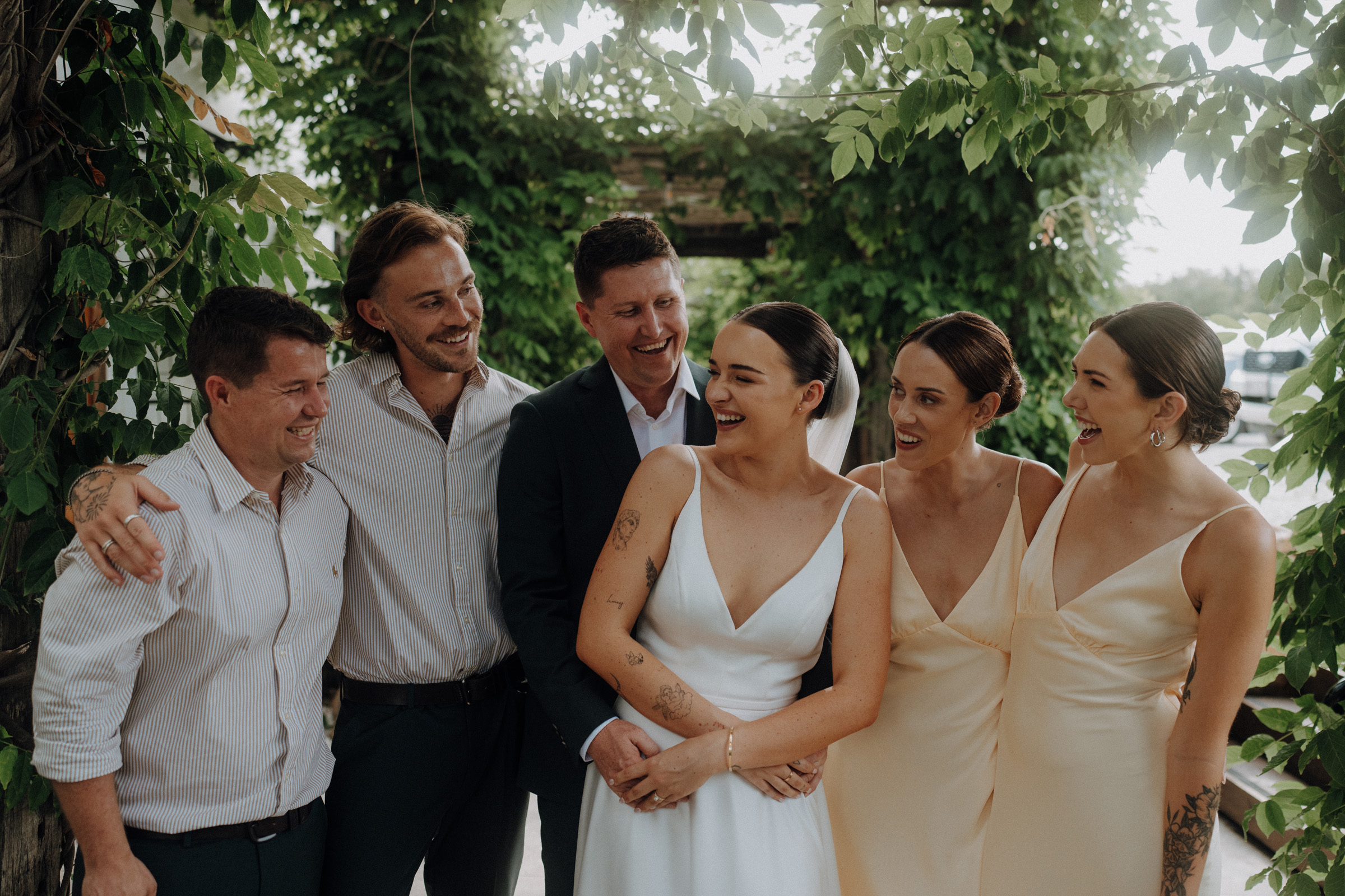 A group of six people, including a bride in a white dress, smiling and standing together outdoors with greenery in the background.