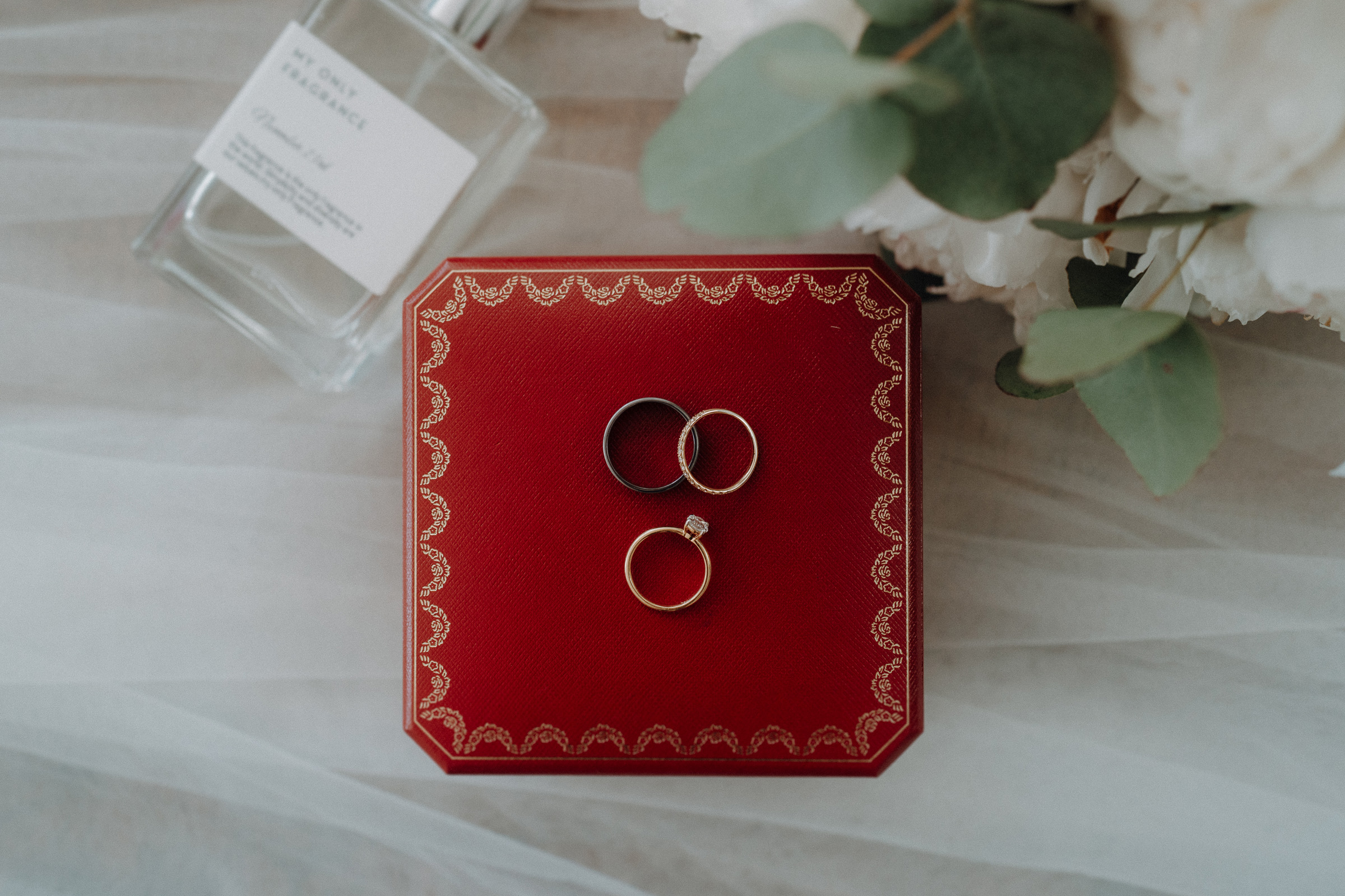 Three rings on a red box next to a perfume bottle and eucalyptus leaves.