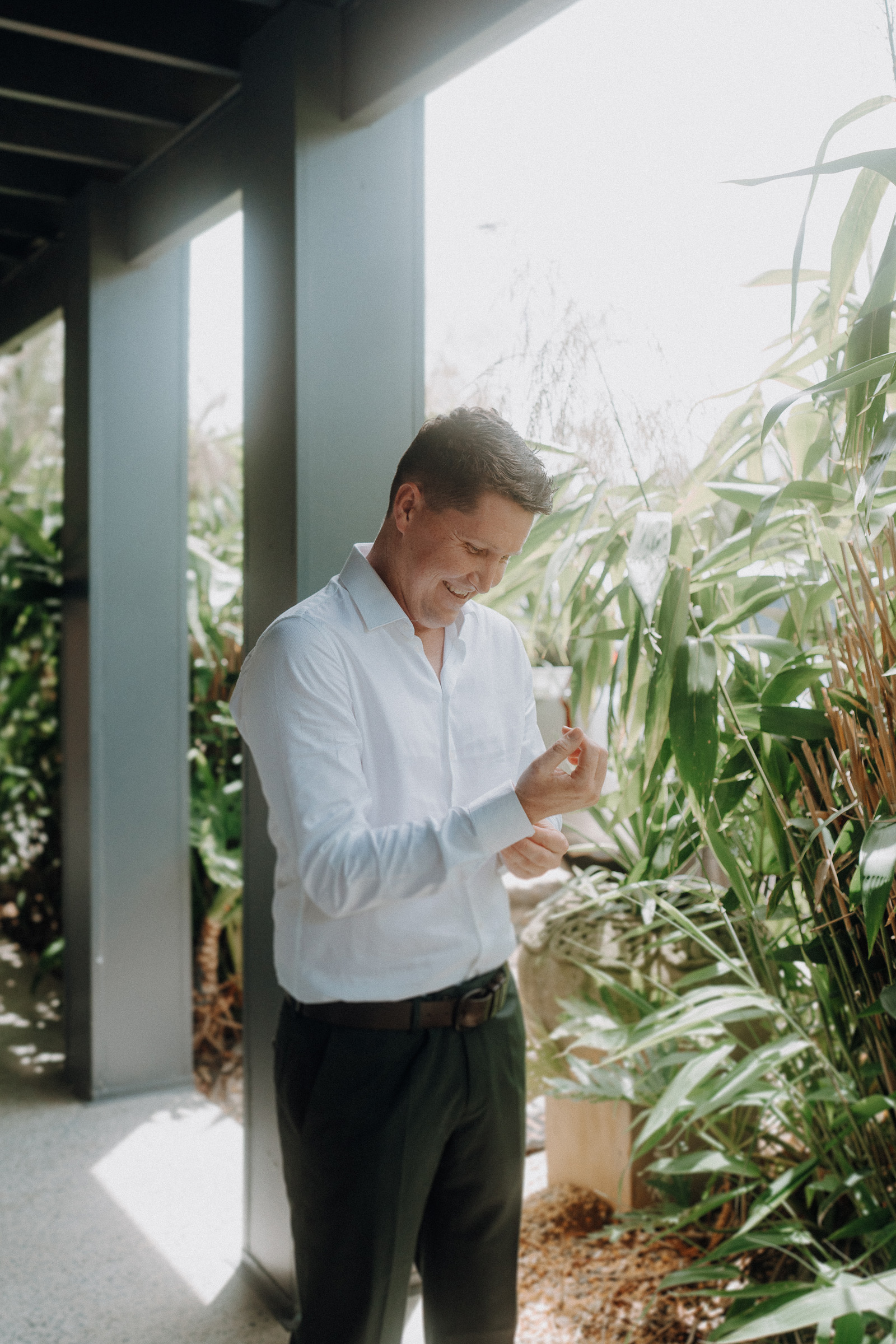 A man in a white shirt stands next to greenery, smiling and looking down at a small object in his hand.