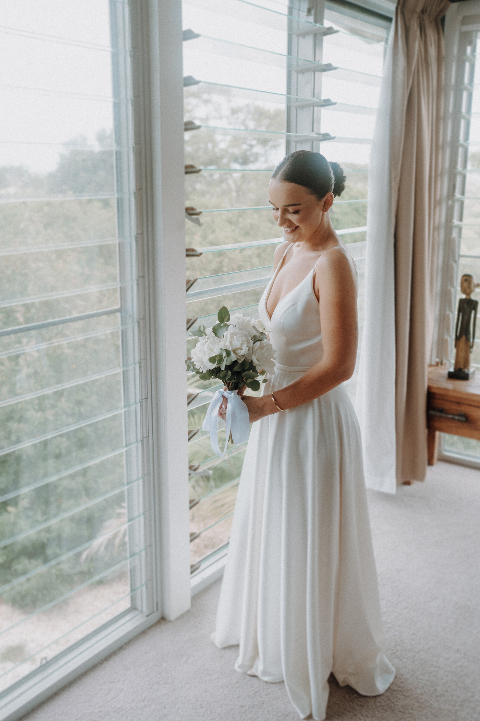 A woman in a white dress stands by tall windows, holding a bouquet of white flowers.