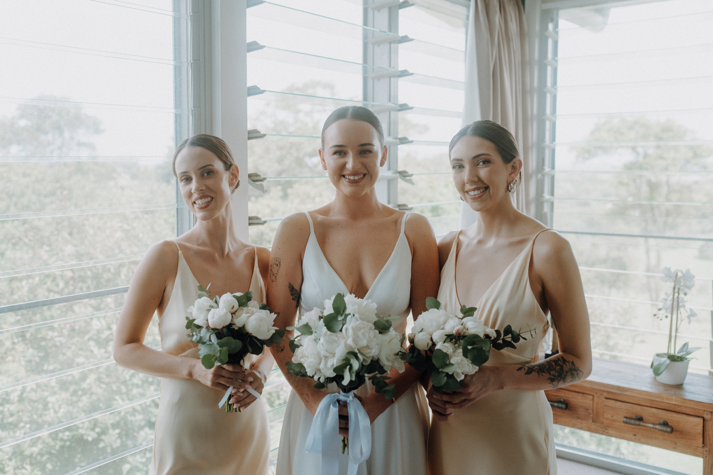 Three people stand indoors by a window, each holding a bouquet of white flowers. They are dressed in light-colored formal attire.