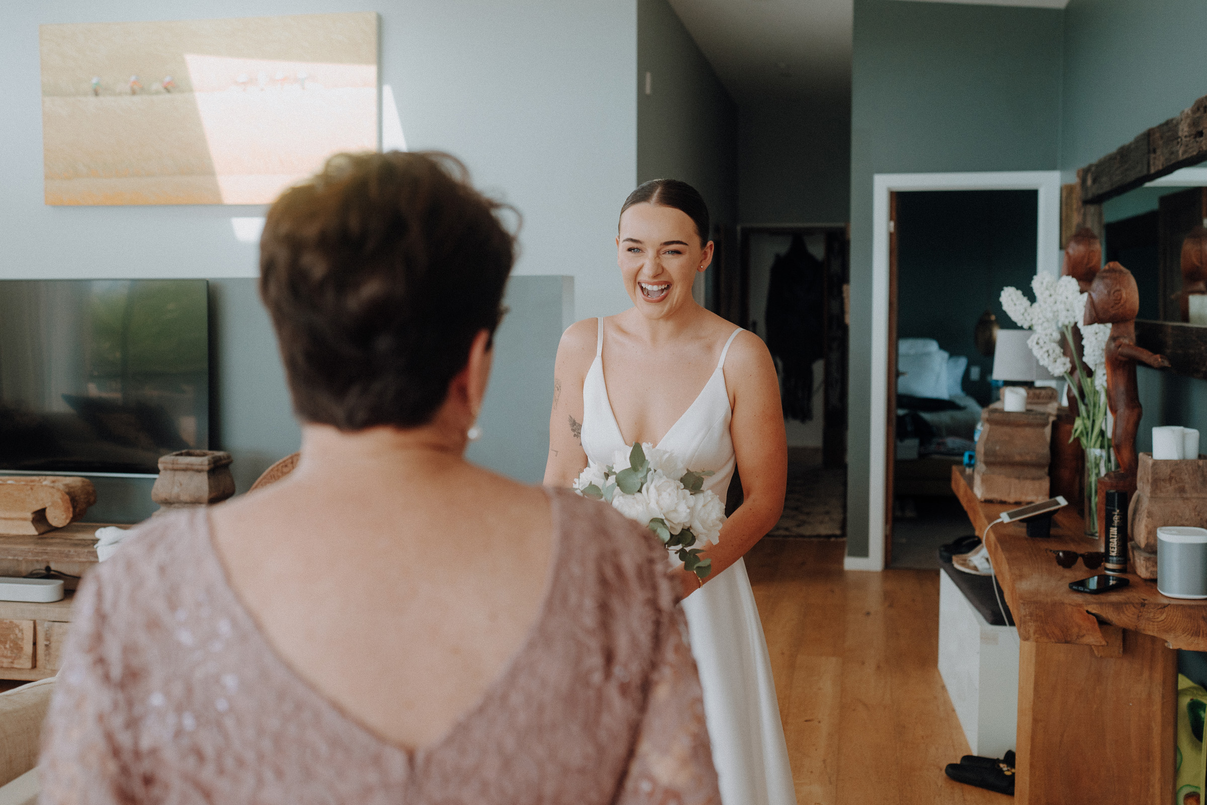 A bride holding a bouquet smiles at a woman in a warmly lit room with modern decor.