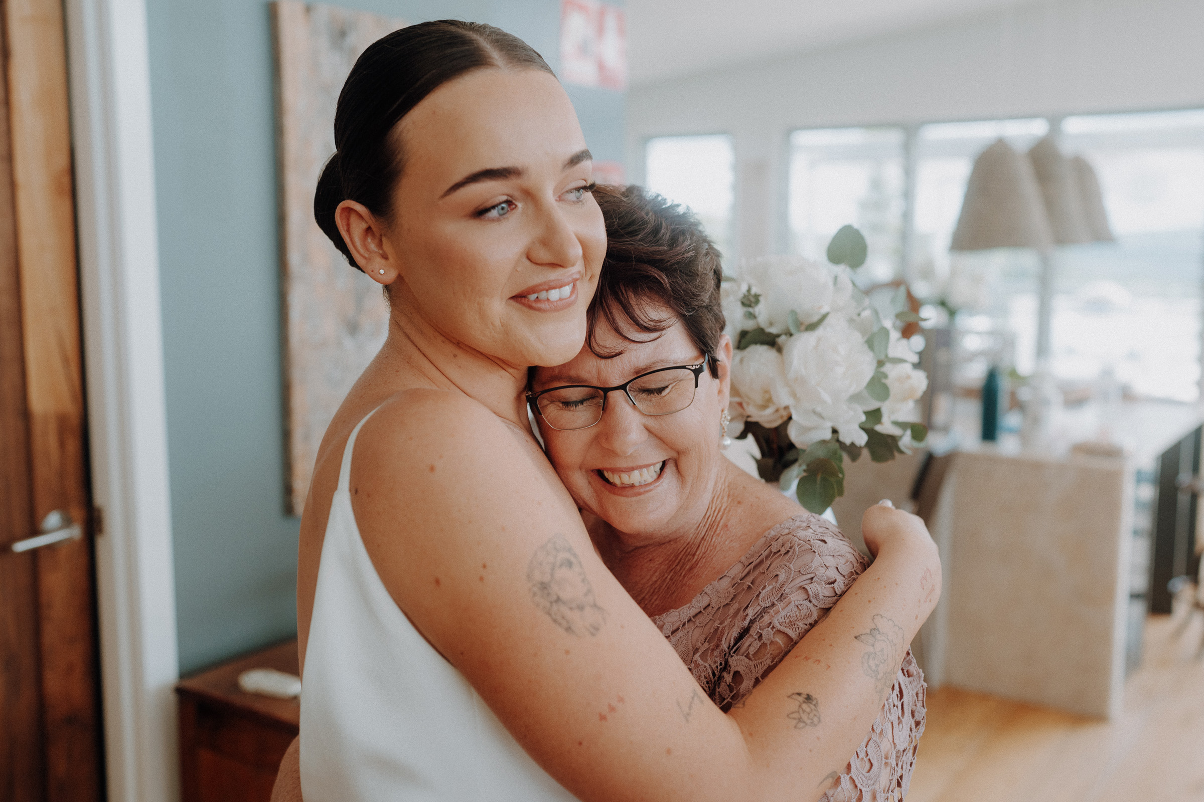 A woman in a white dress hugs a smiling woman wearing glasses, holding a bouquet of white flowers in a bright room.