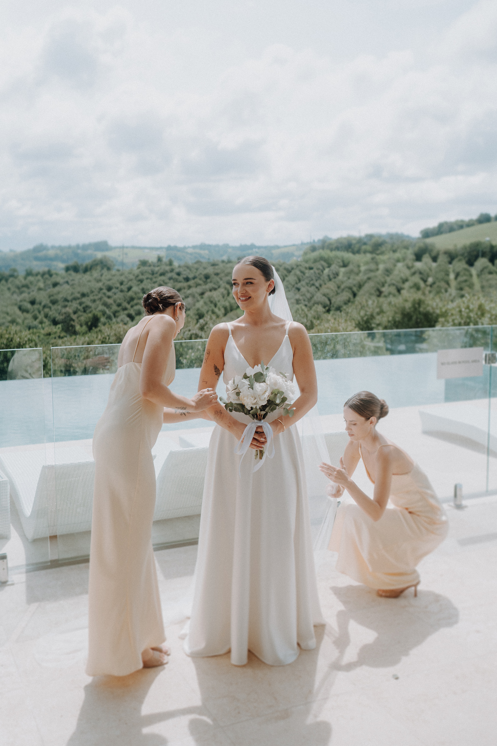 Bride holding flowers stands on a terrace with a scenic view, while two bridesmaids adjust her dress.