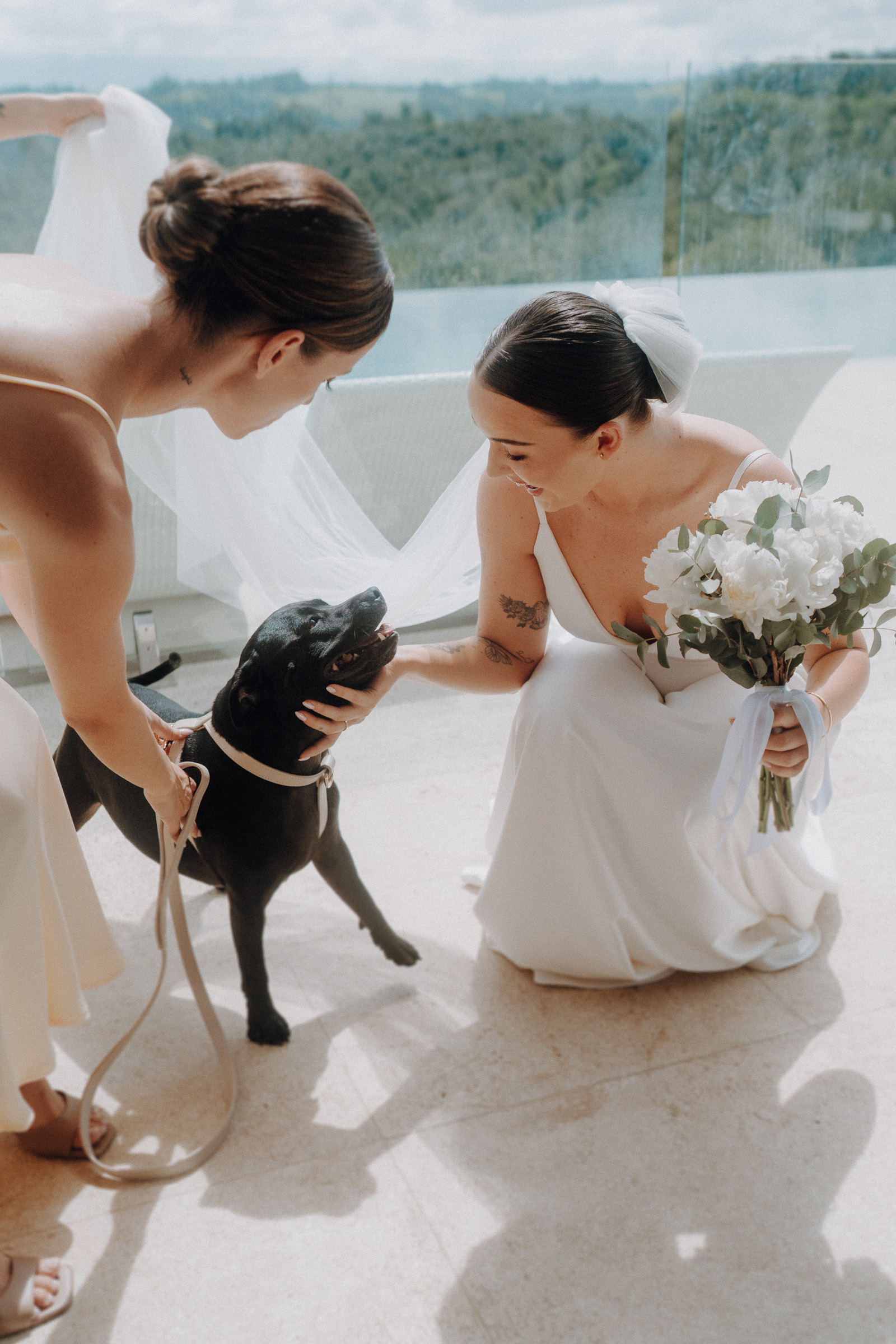 A bride in a white dress holds a bouquet and pets a black dog, while another woman in a light dress holds the dog's leash outdoors.