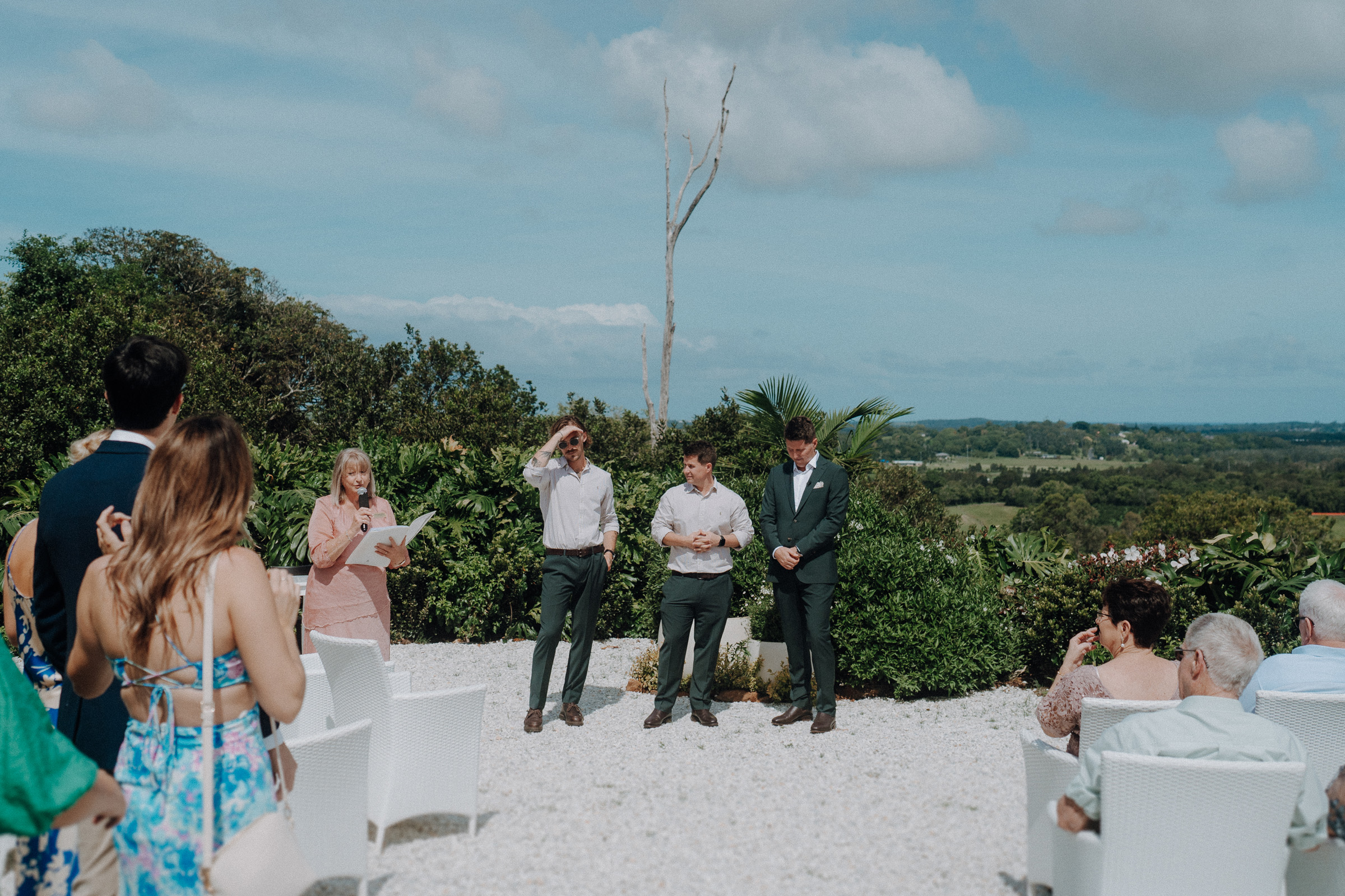 Outdoor wedding ceremony with guests seated, a person reading from a paper, and three people standing in front of lush greenery and a distant landscape.