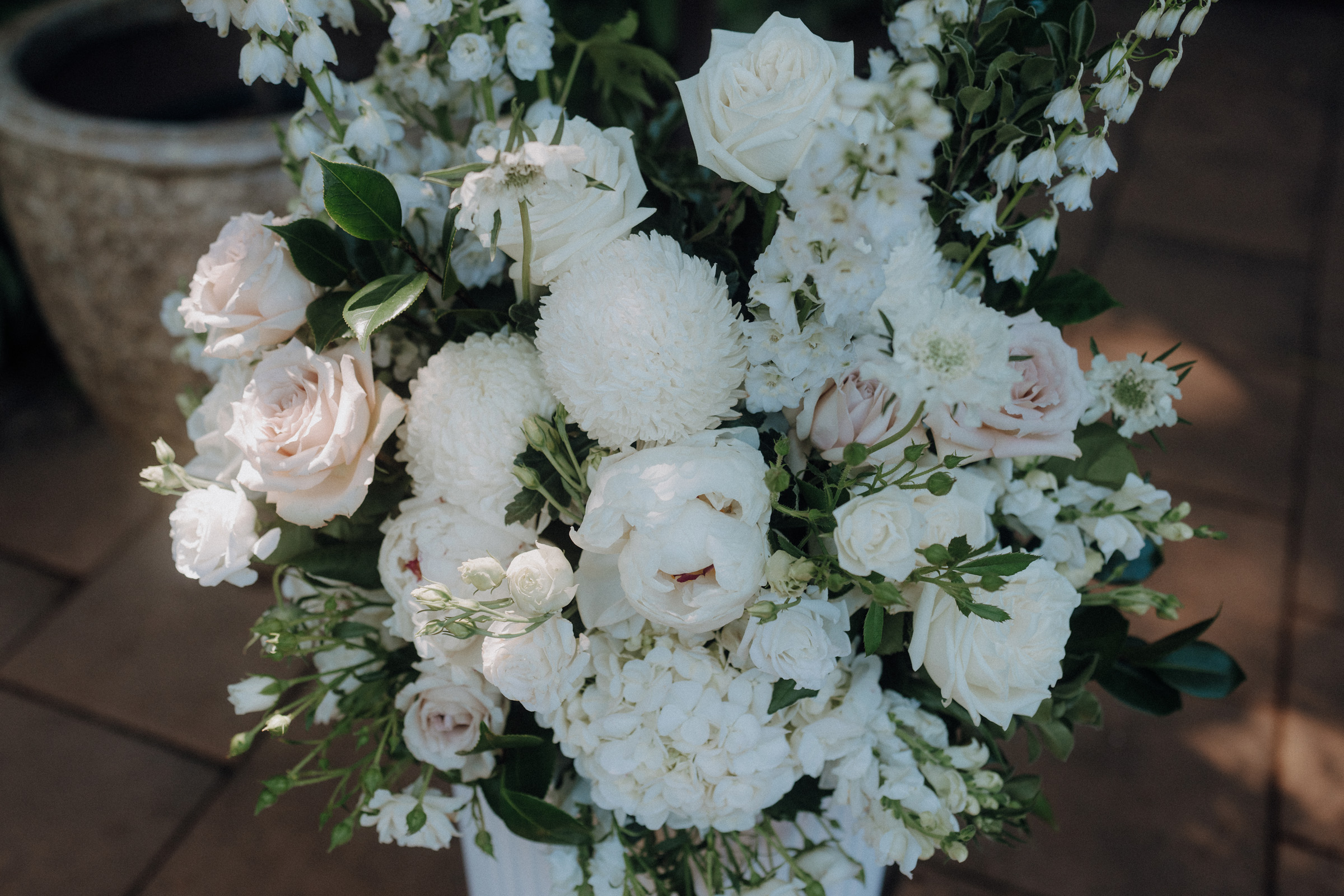 A floral arrangement featuring white and pale pink roses, chrysanthemum blooms, and assorted greenery on a stone surface.