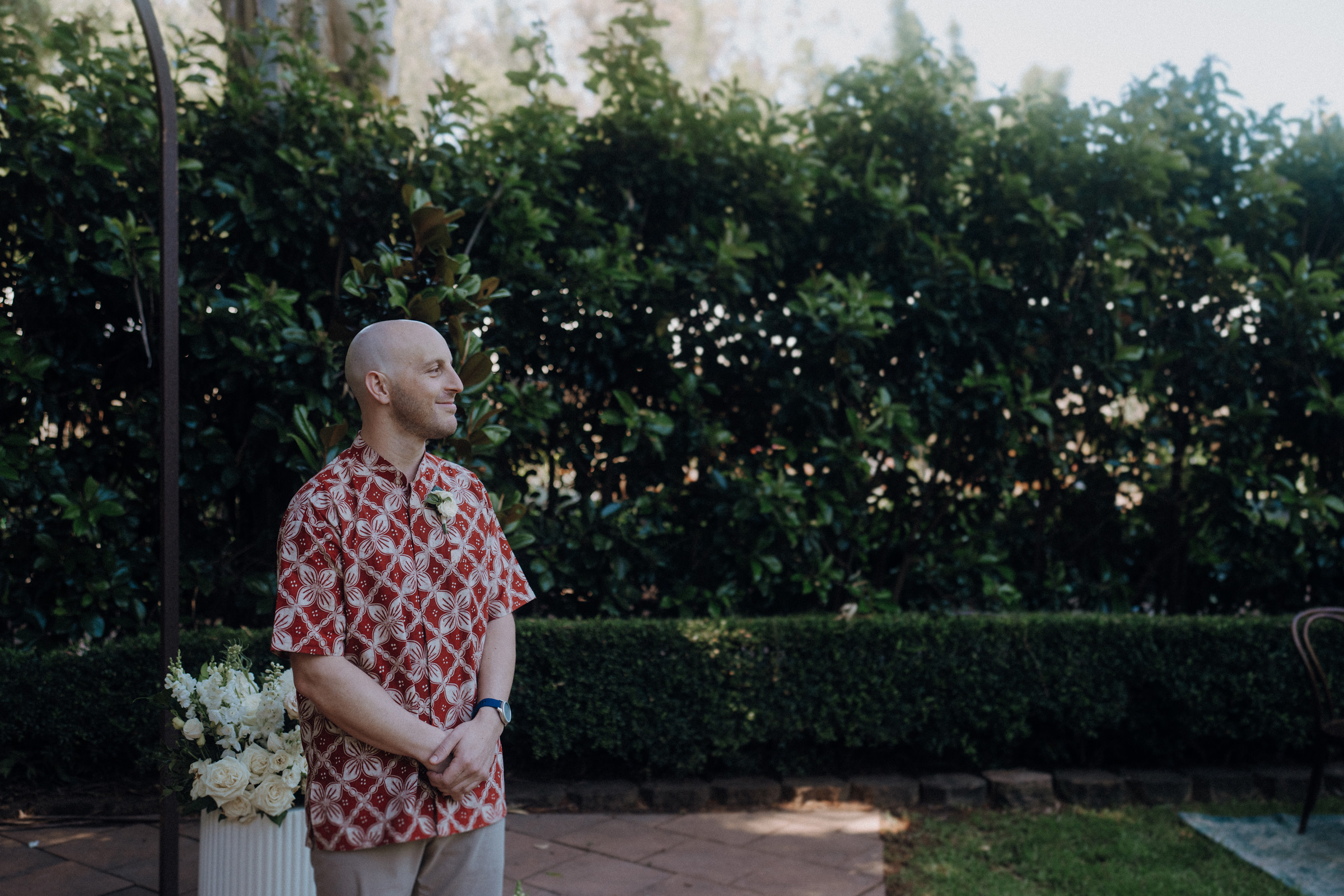 A person stands outdoors on a paved path wearing a patterned shirt, with bushes and greenery in the background.