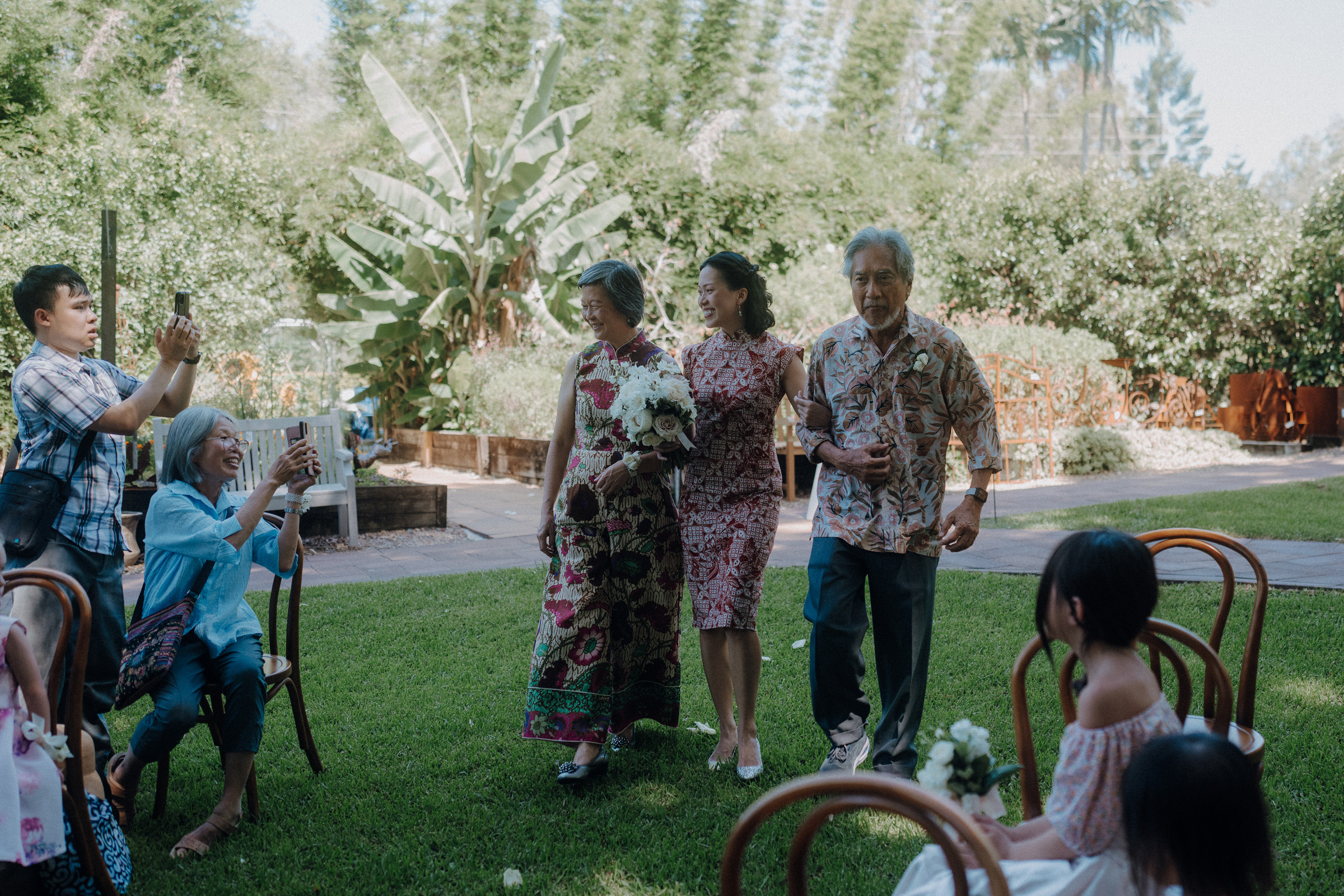 An elderly couple is being assisted down a grassy aisle by a younger woman. People seated nearby are taking photos. The background is lush with greenery.