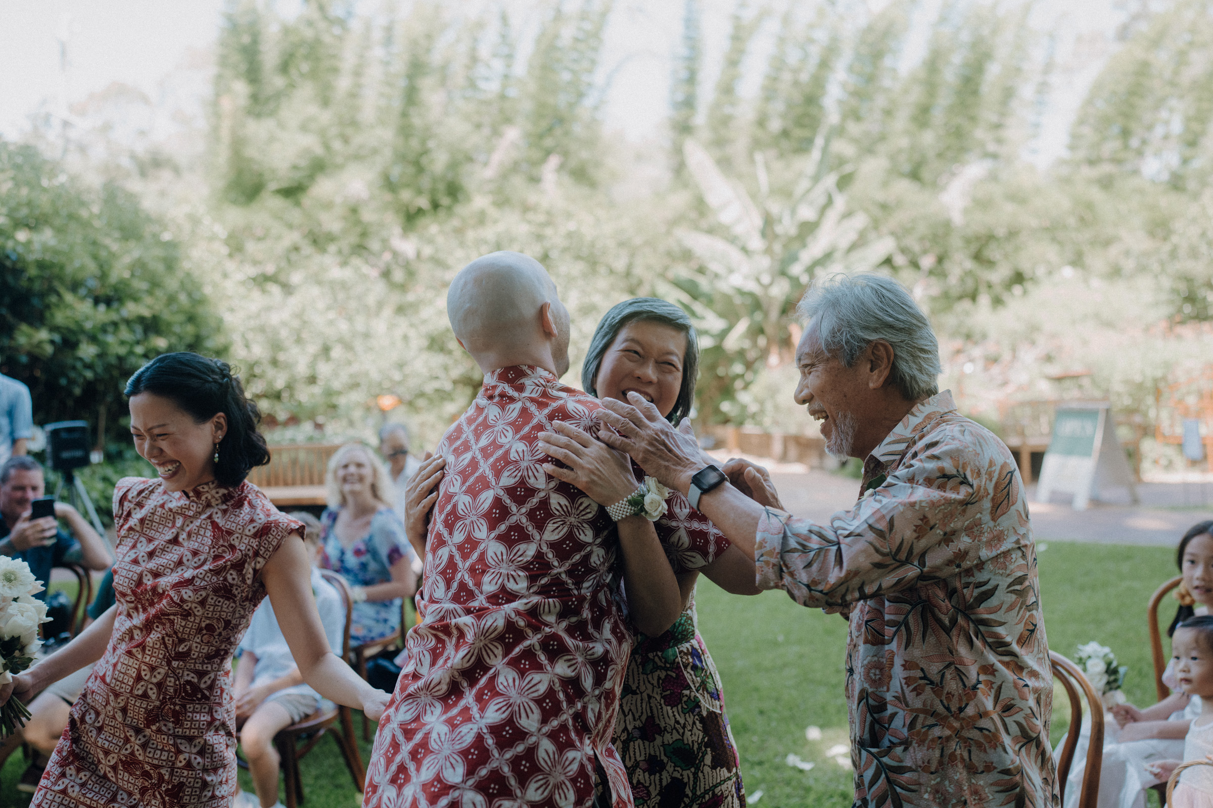 A group of people in patterned clothing hug and laugh outdoors on a grassy area, surrounded by seated onlookers and lush greenery.