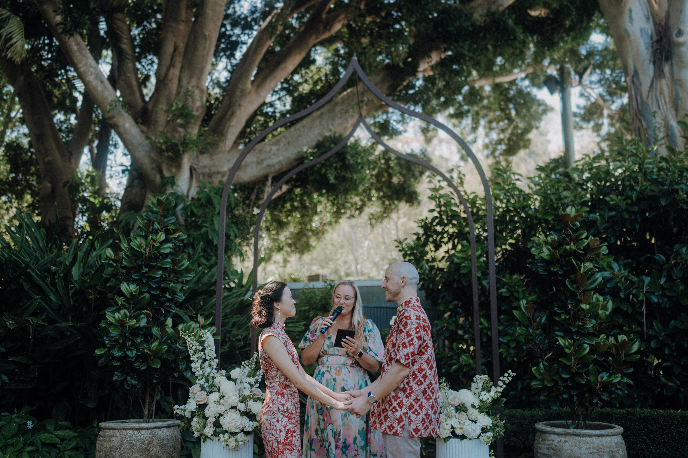 A couple holds hands during an outdoor wedding ceremony. A person stands in front of them, reading. The setting includes lush greenery and floral arrangements.