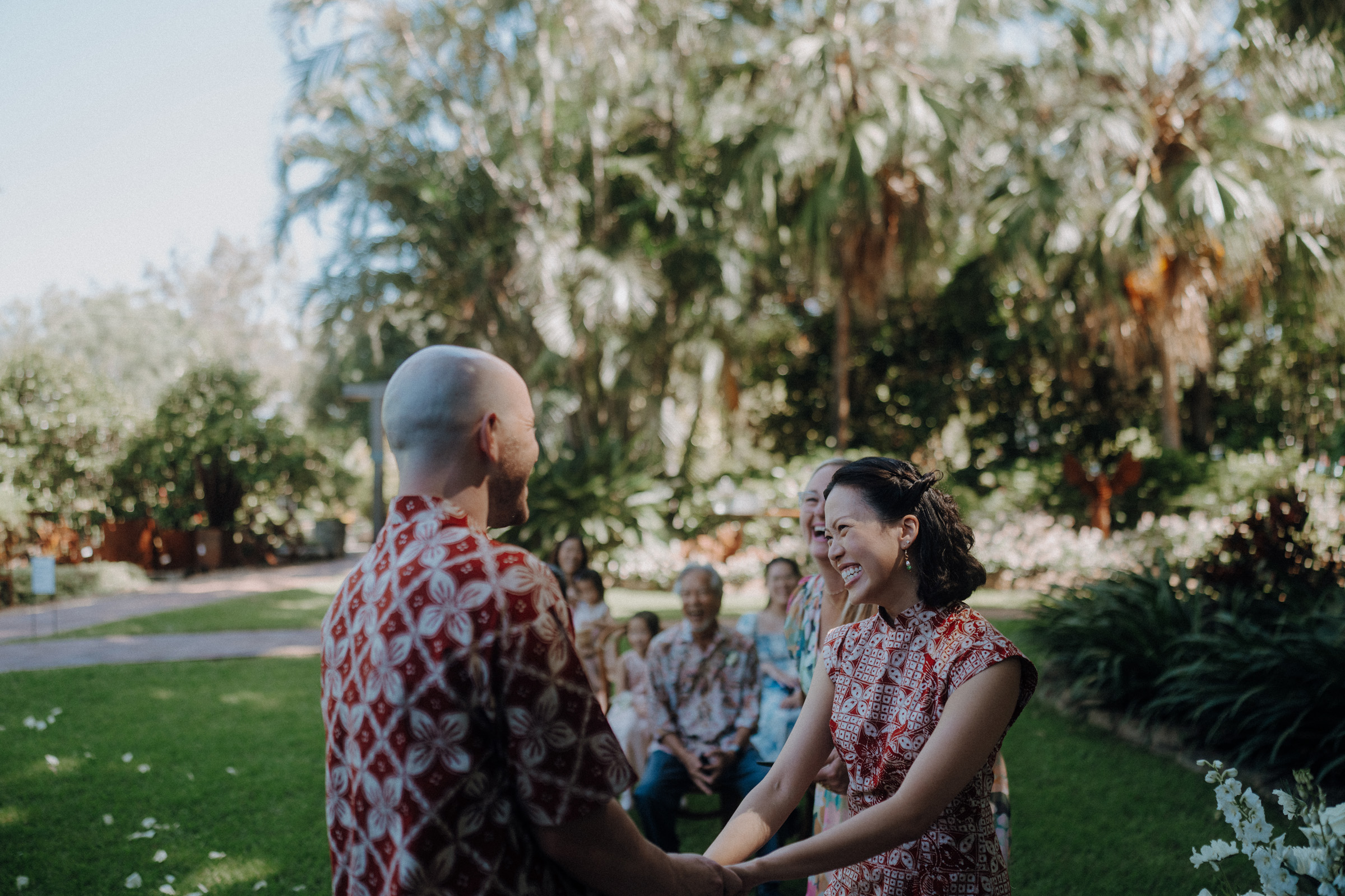 A couple holds hands in a garden ceremony, surrounded by a few seated guests. They are wearing red patterned outfits, and lush greenery is visible in the background.