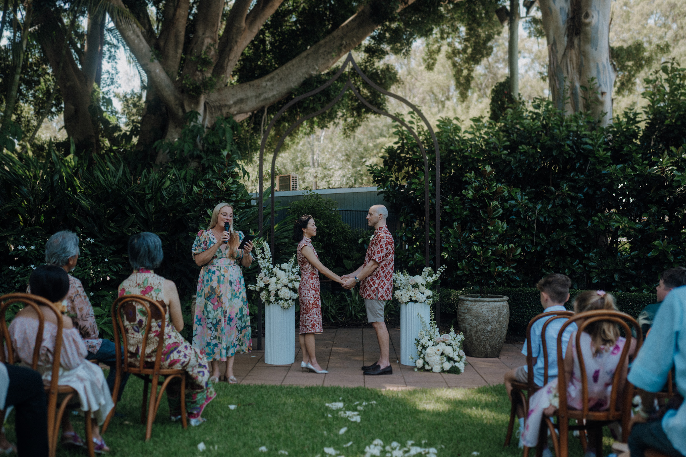 A couple stands holding hands during an outdoor ceremony, with a person officiating. Guests are seated on either side, surrounded by greenery and floral arrangements.