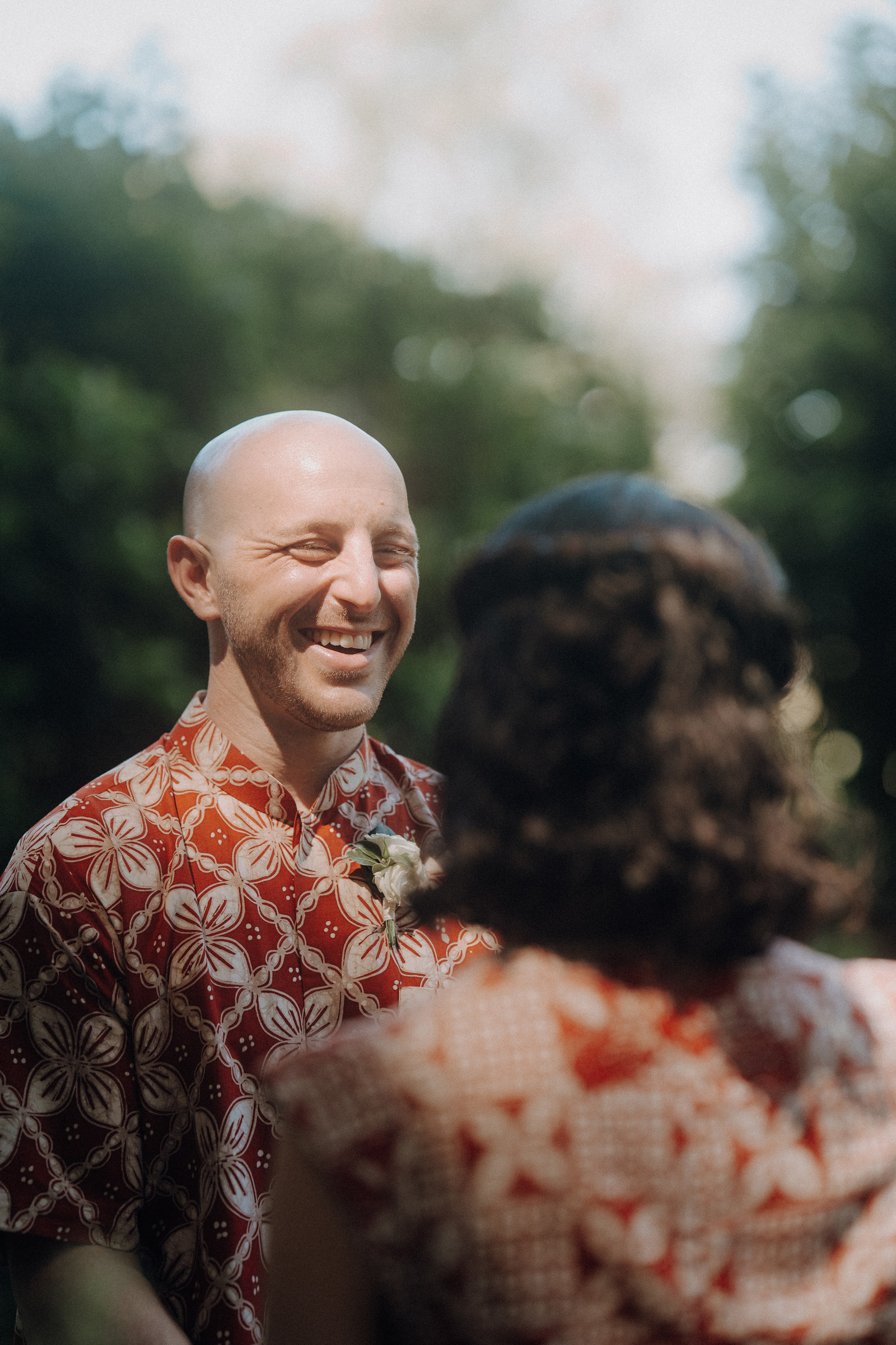 A bald man wearing a red patterned shirt smiles at a woman with curly hair outdoors in a lush green setting.