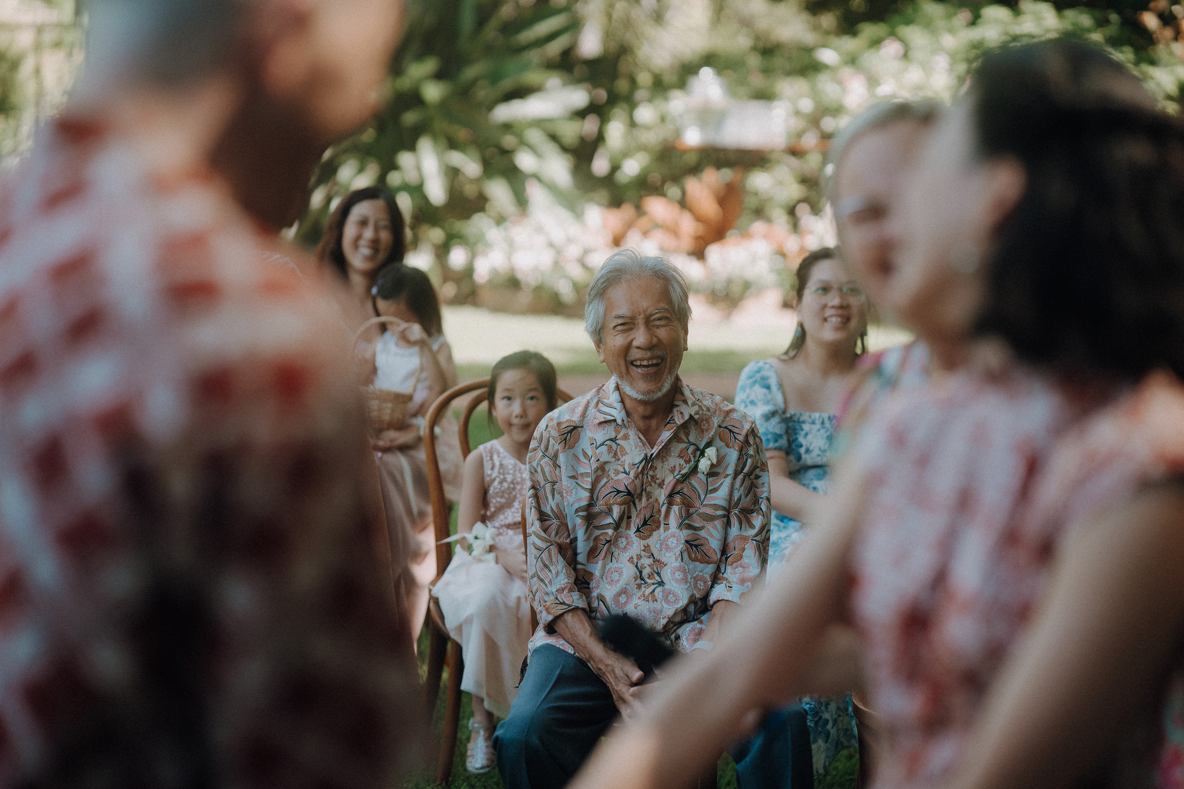 A group of people, including children and adults, are seated and smiling outdoors, surrounded by greenery. One man at the center is looking forward with a cheerful expression.