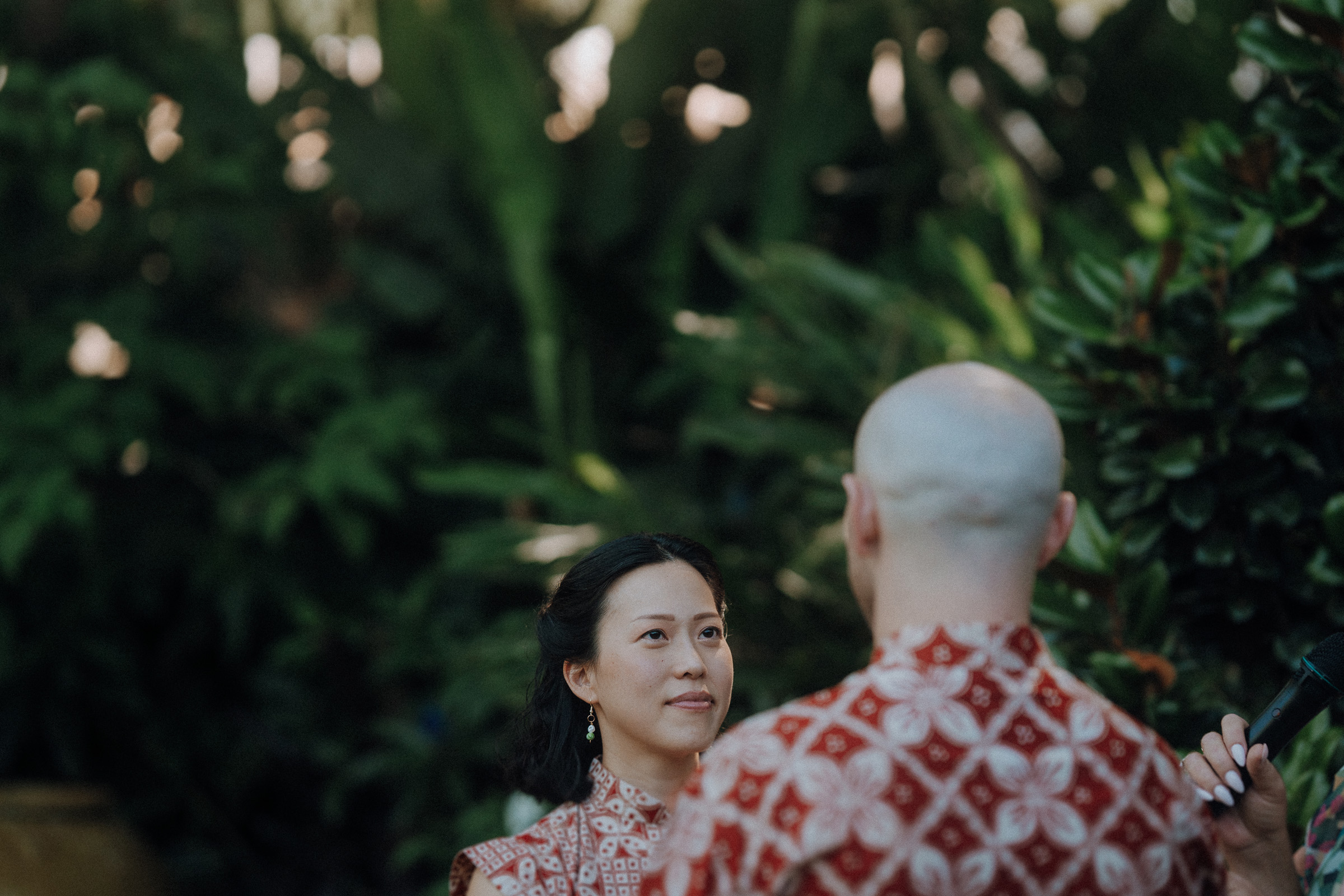 A person with long hair and another bald person, both in red-patterned clothing, stand outdoors surrounded by greenery.