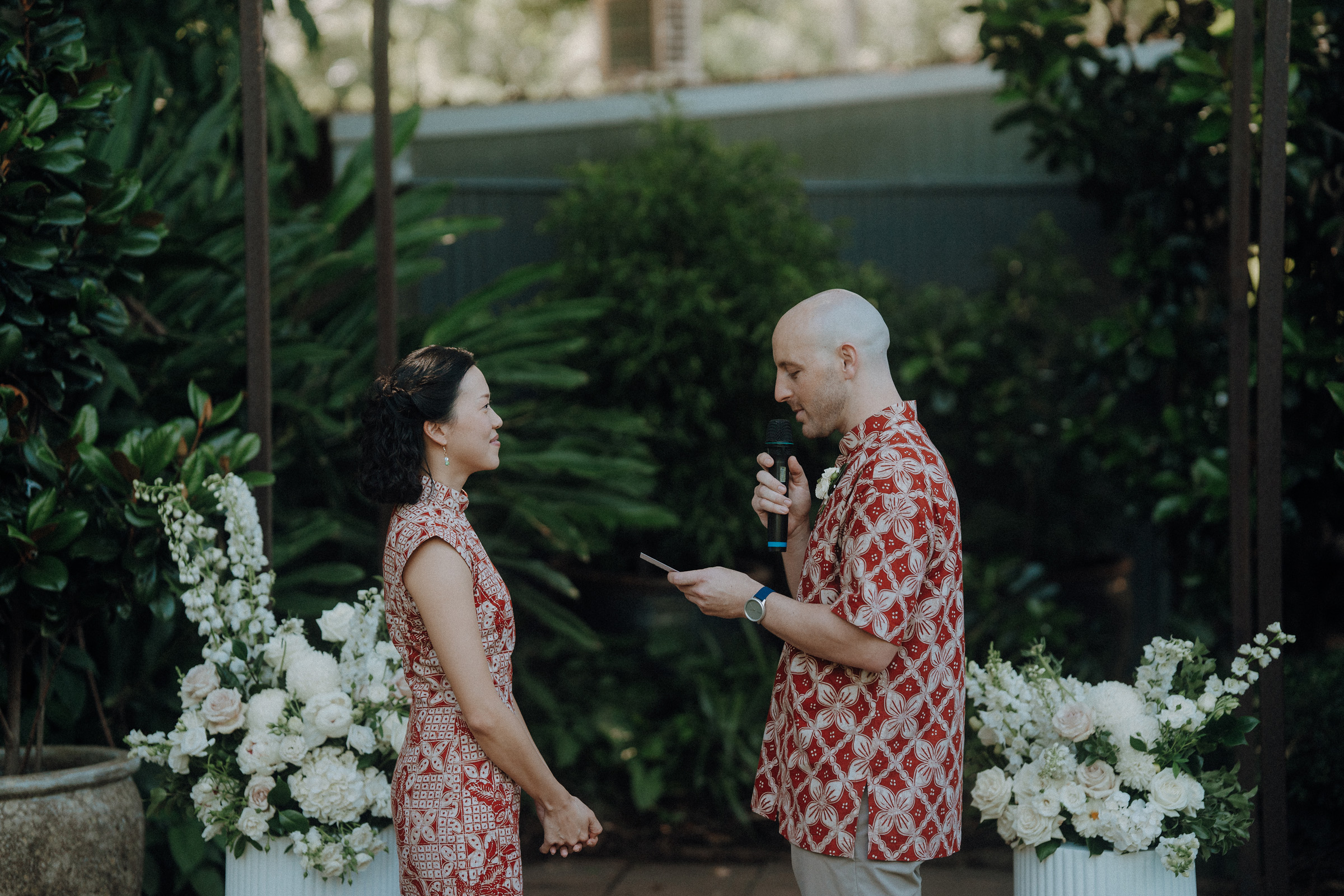 A couple in matching red patterned outfits stand facing each other during a ceremony in a lush garden setting, surrounded by white floral arrangements.