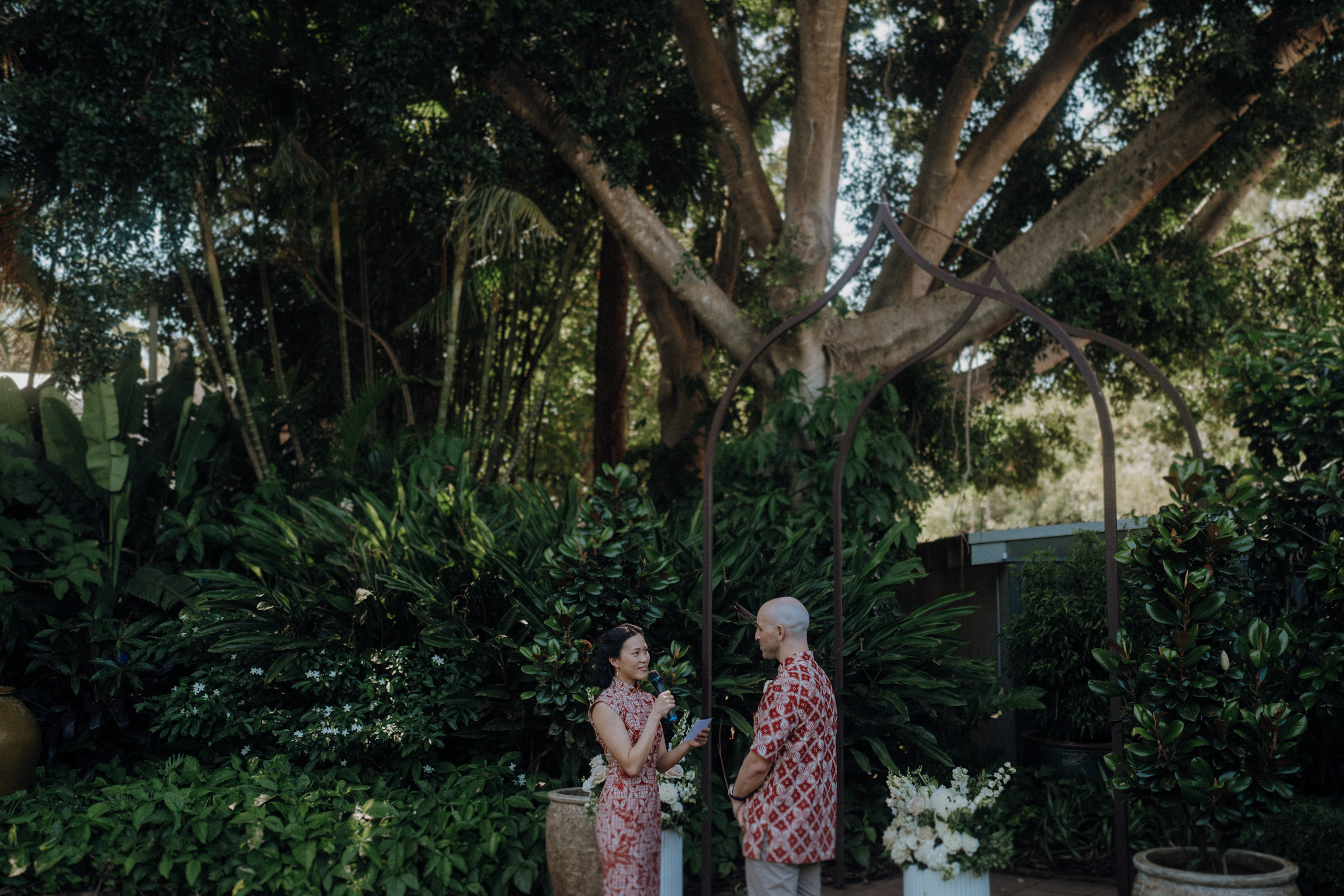 A couple stands together under a decorative arch, surrounded by lush greenery and large trees.