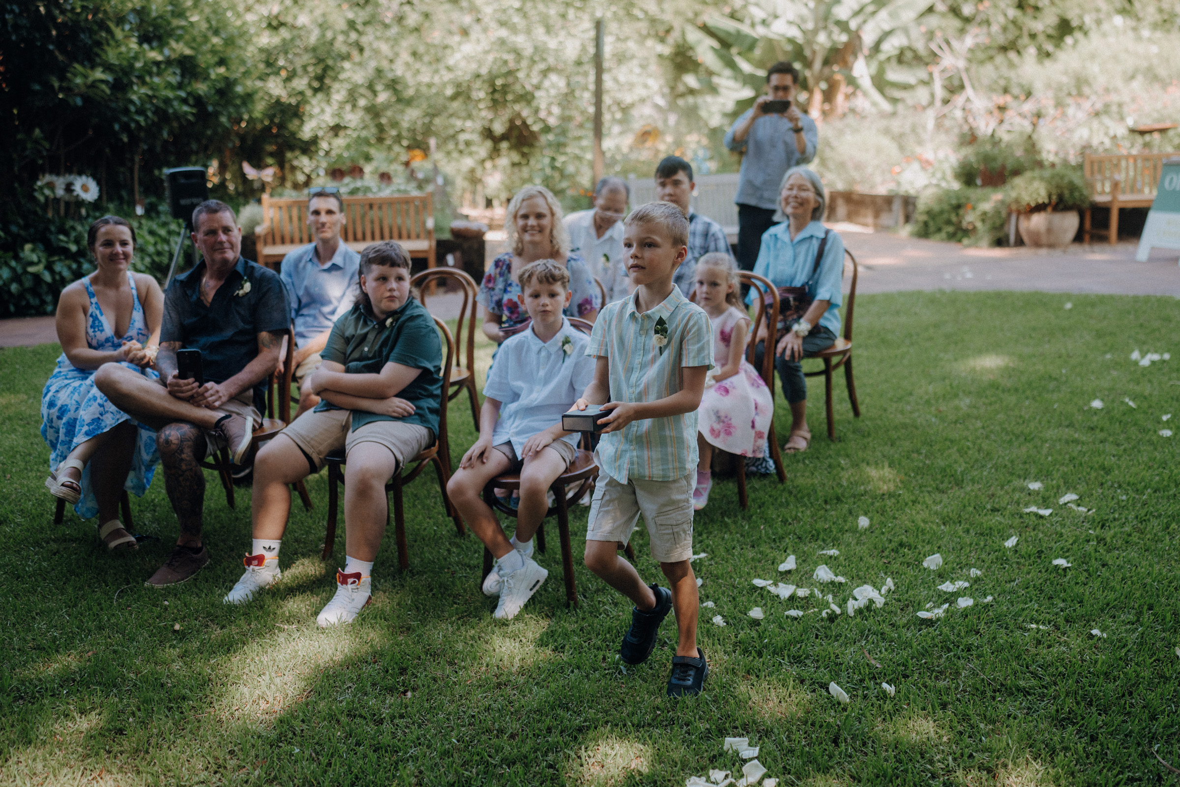 A boy walks on grass holding a small box, while a seated group of adults and children watch. Trees and foliage are in the background.