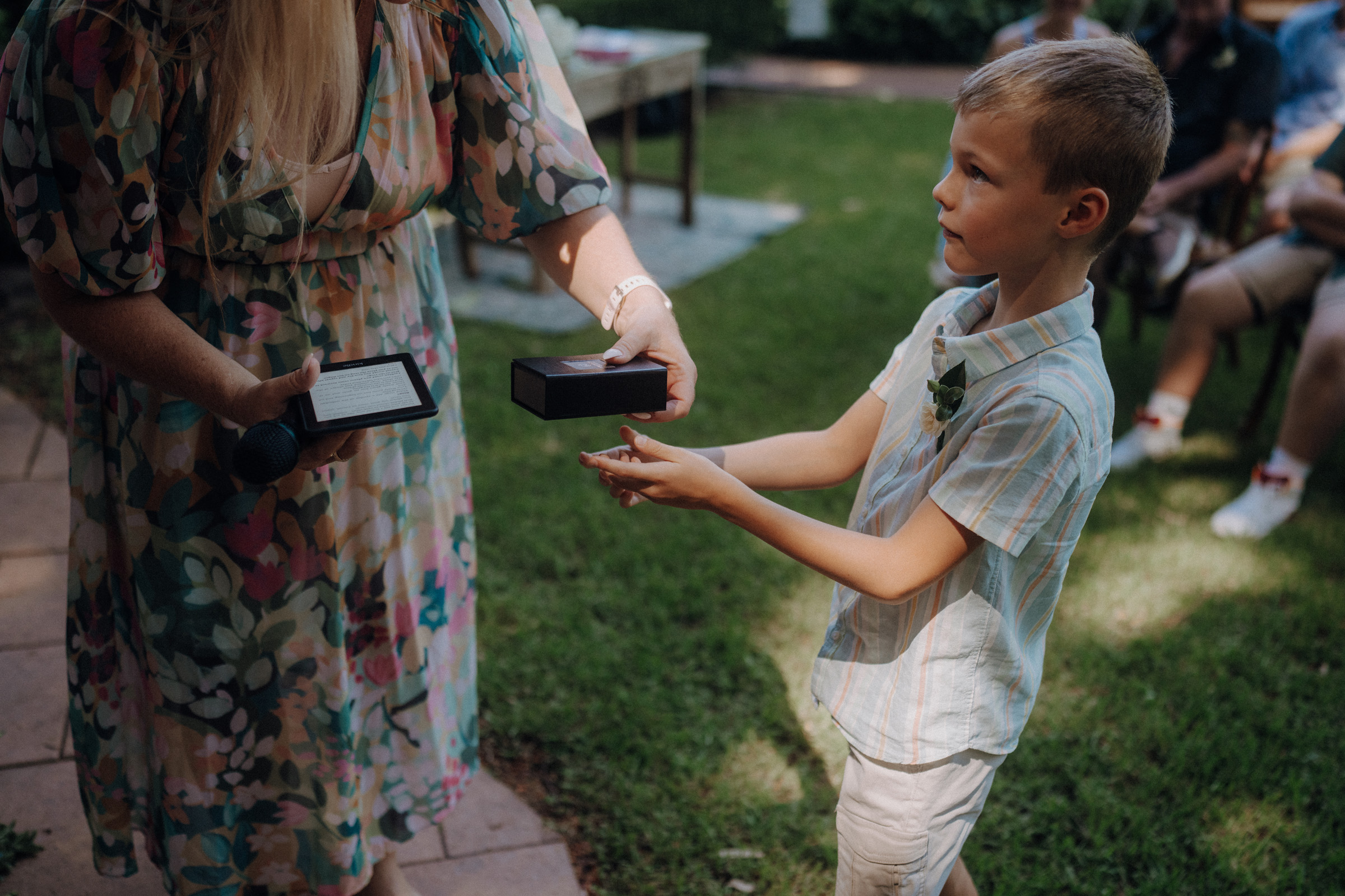 A young boy in a striped shirt receives a small box from a woman in a floral dress during an outdoor gathering.