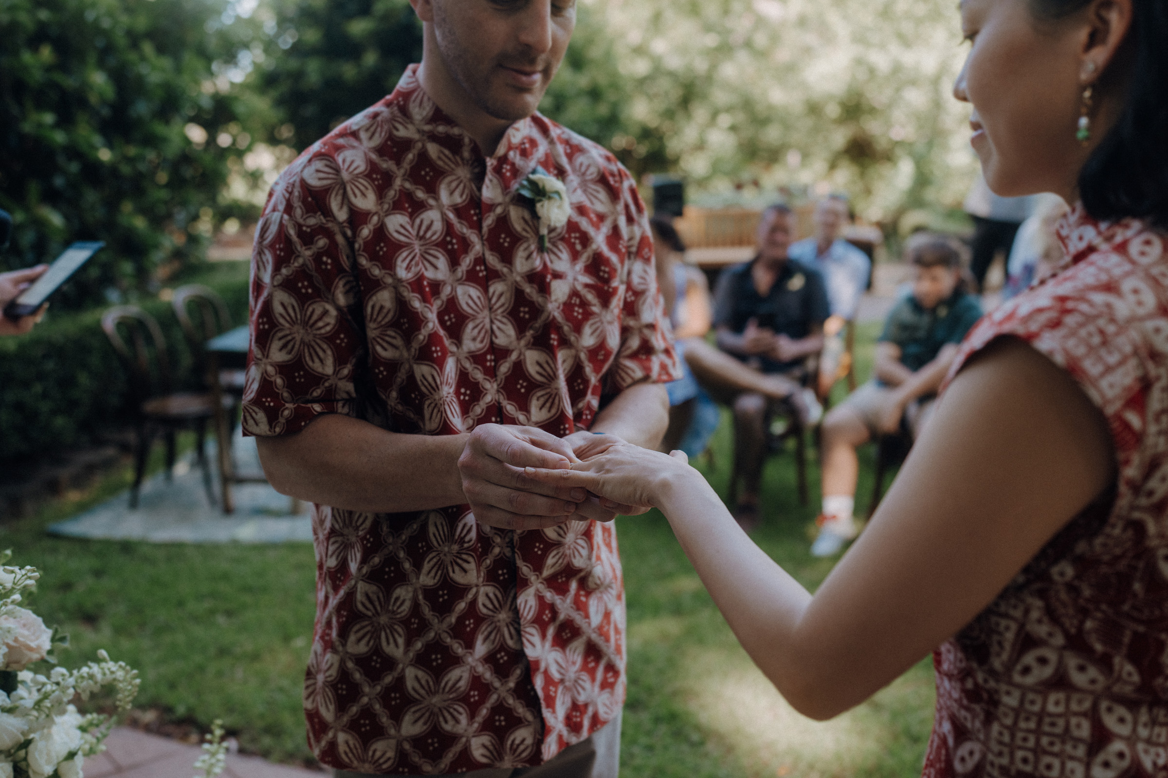 A couple exchanges rings during an outdoor ceremony. The man wears a patterned shirt while the woman holds out her hand. Guests are seated in the background.