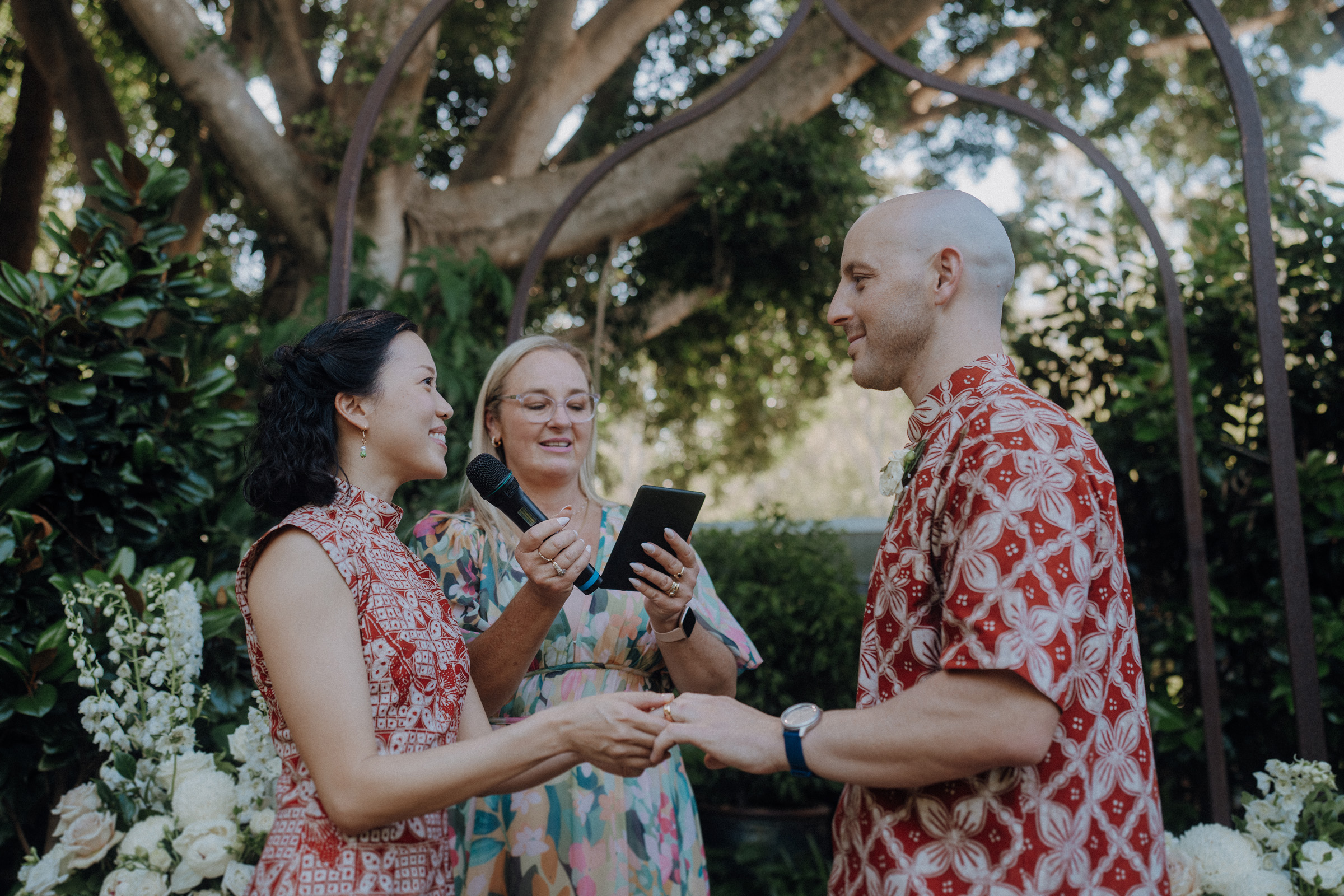 A couple in red patterned attire exchange rings during an outdoor ceremony, with an officiant reading from a book, surrounded by greenery and floral decorations.