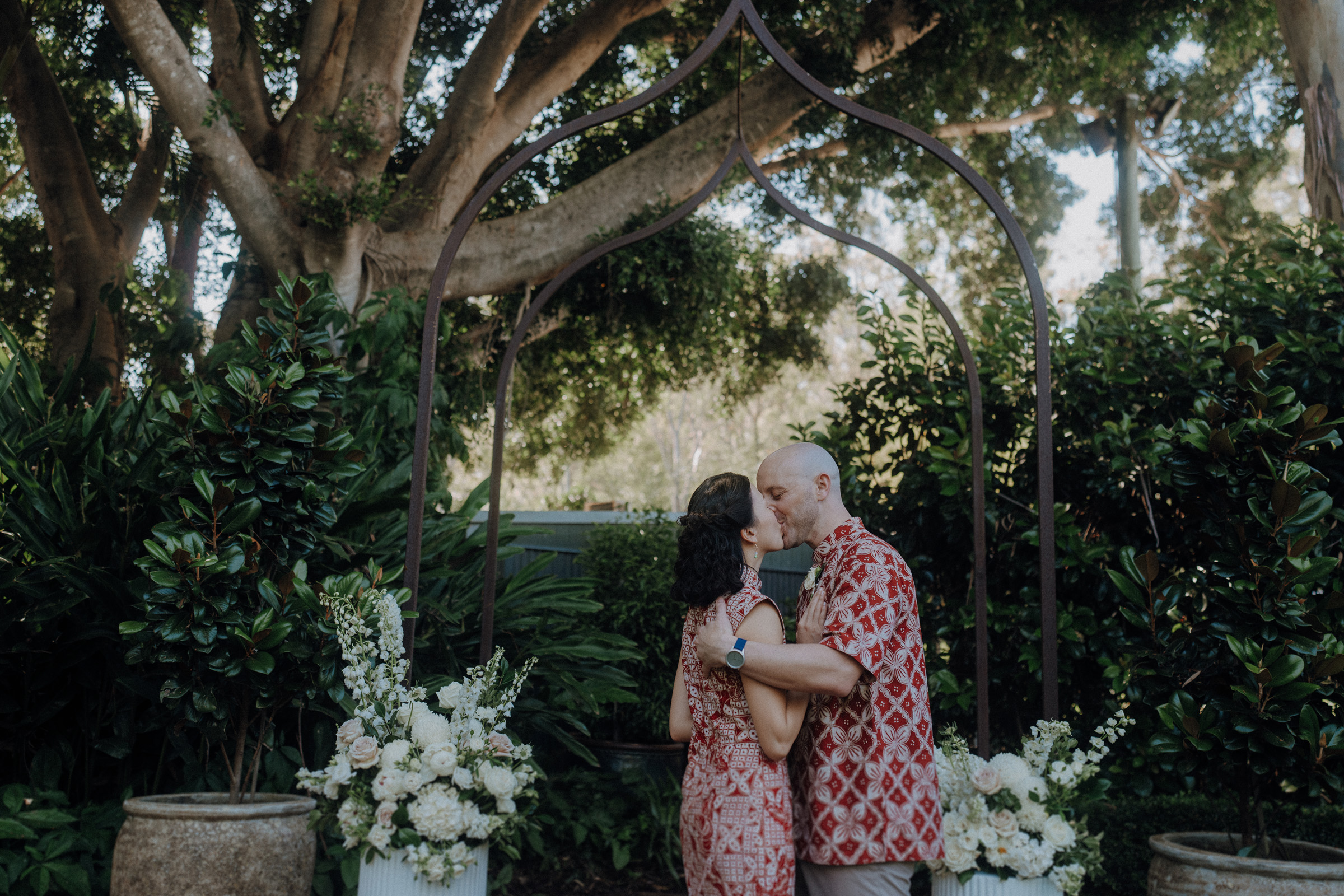 A couple in patterned outfits kisses under a decorative arch surrounded by lush greenery and white floral arrangements.