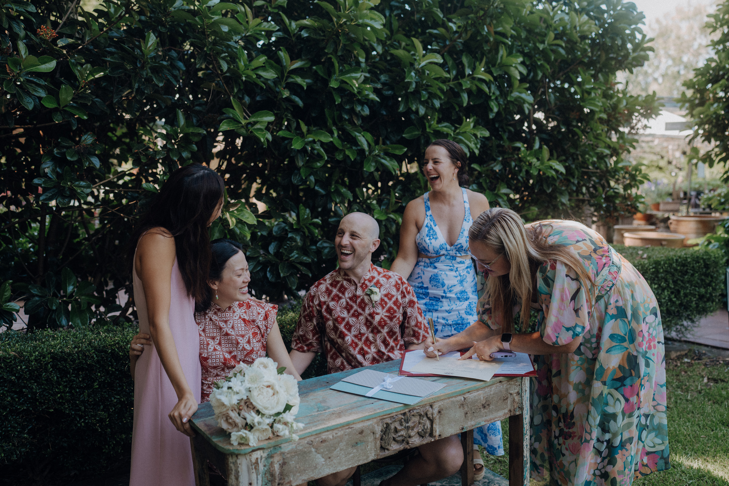 A group of people in colorful attire gather around a rustic table outdoors, with some smiling and one person writing. A bouquet of flowers rests on the table.