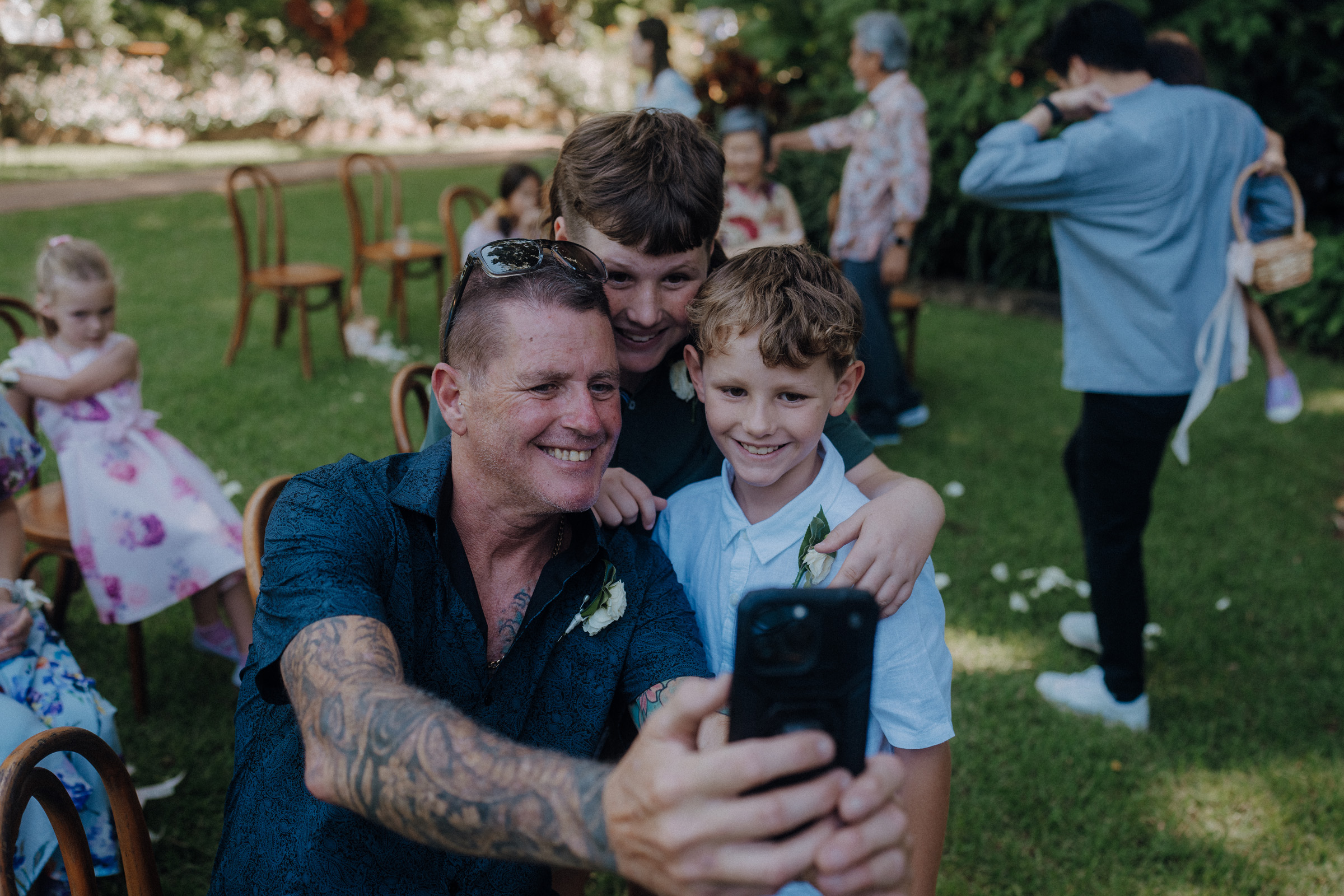 A man with tattoos takes a selfie with two smiling boys outdoors. Other people and children are in the background.