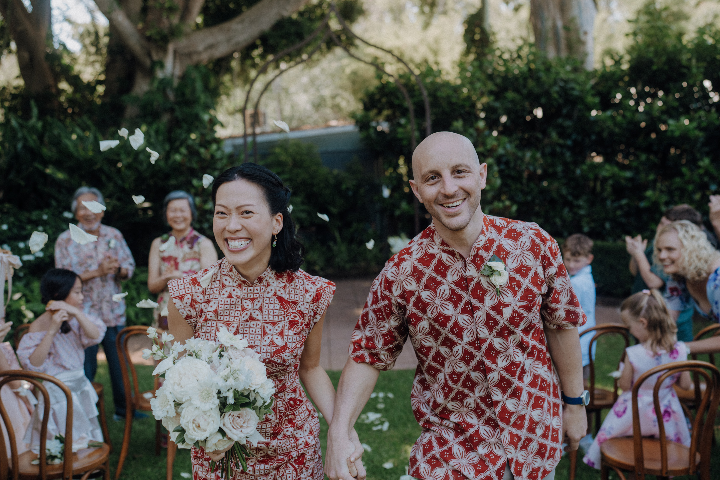 A couple in matching red patterned attire walks down an outdoor aisle, smiling and holding hands, with guests and trees in the background.