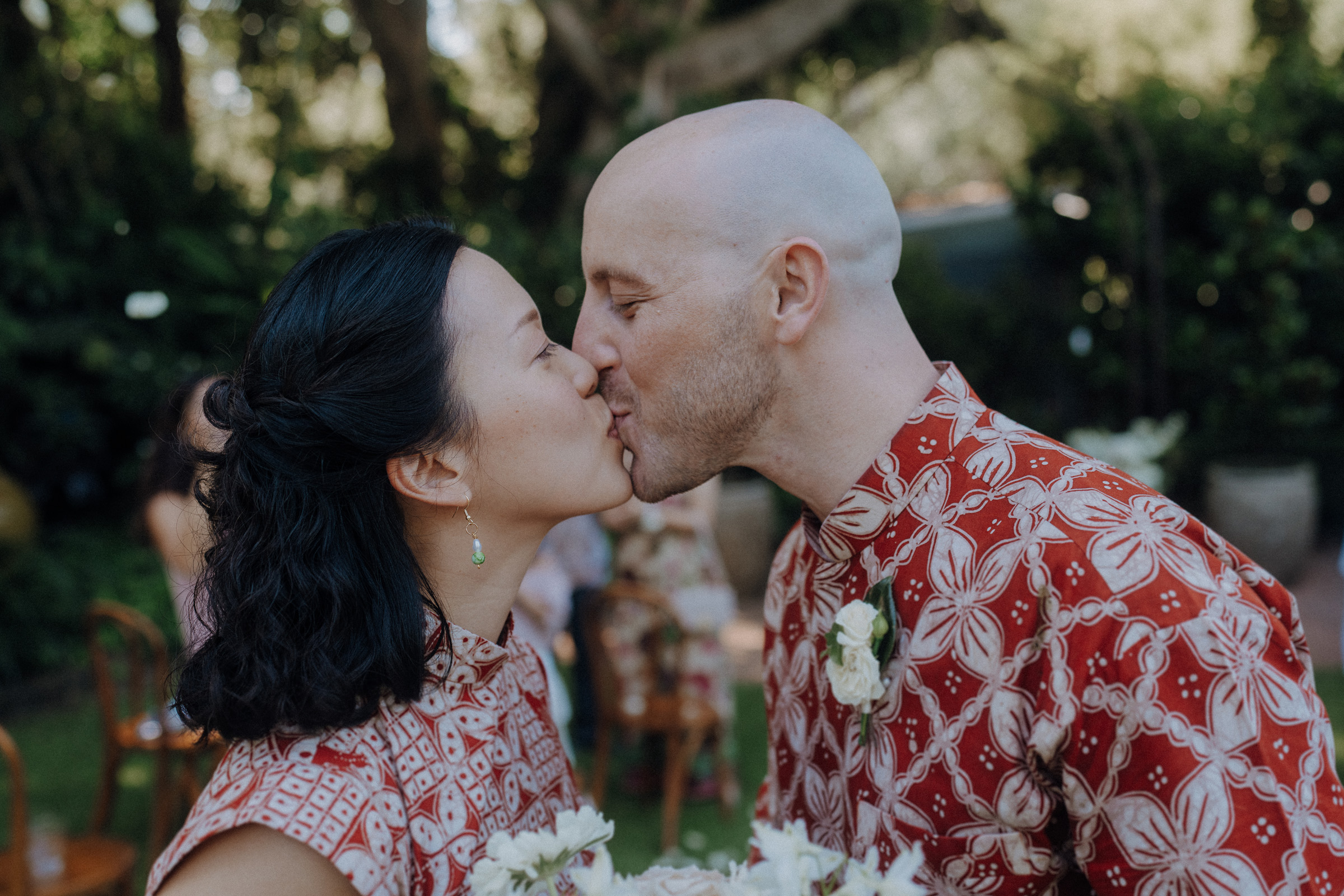 A couple kisses outdoors, both wearing red patterned outfits. They hold flowers and are surrounded by greenery.