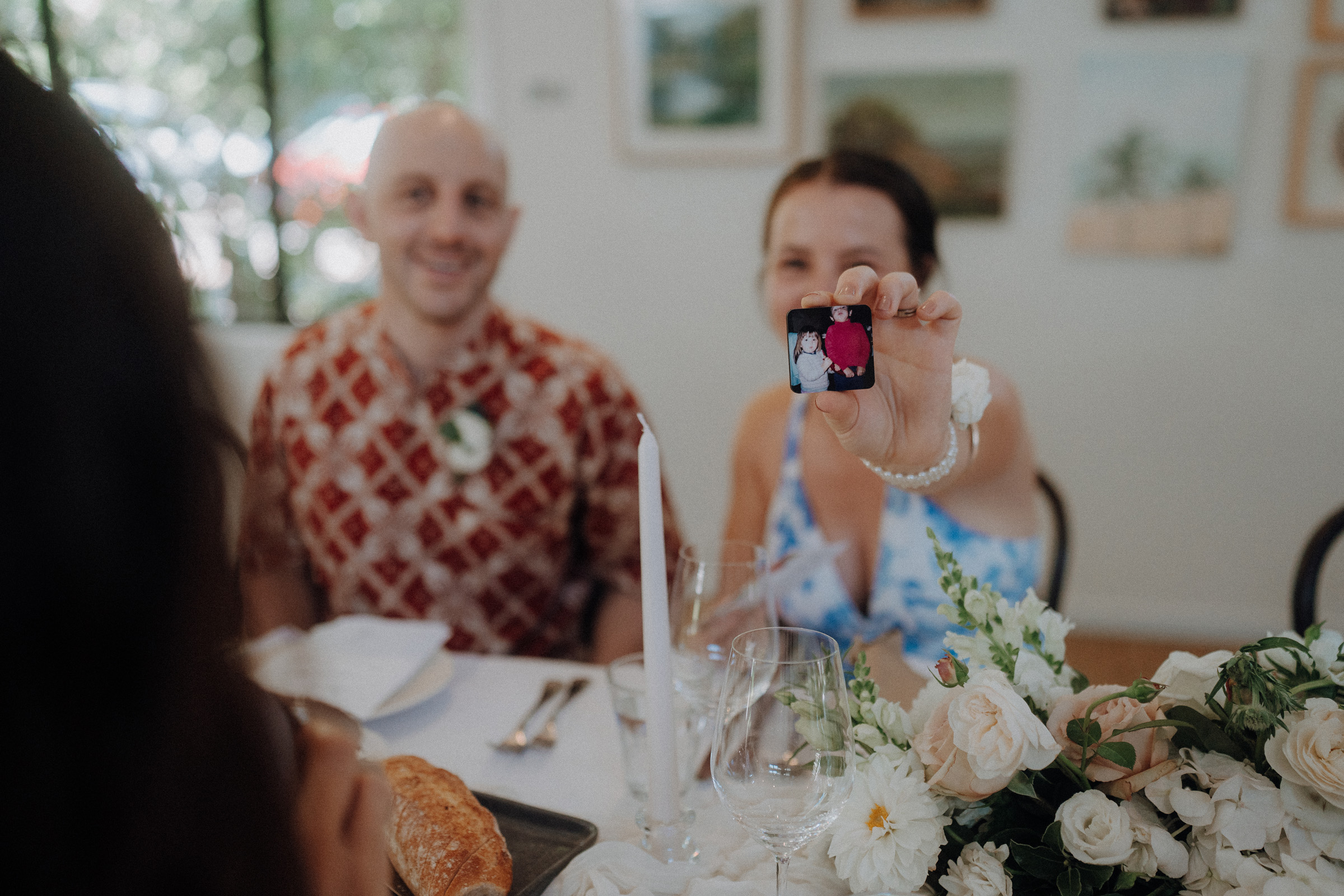 A person holds up a small object with two images on it at a decorated dining table, while another person smiles beside them. Flowers and a loaf of bread are on the table.