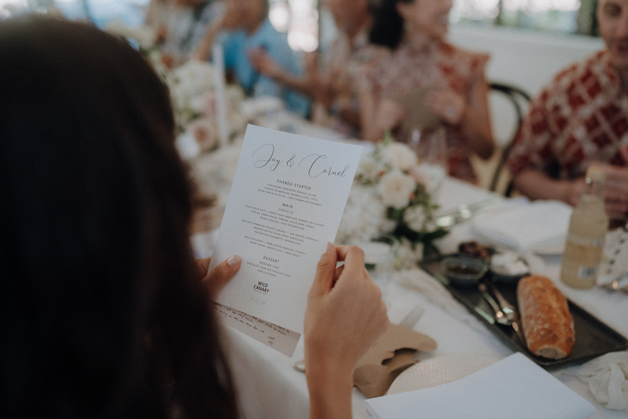 Person holding a menu at a table set for a meal, with a loaf of bread and flowers visible, while others are seated around the table.