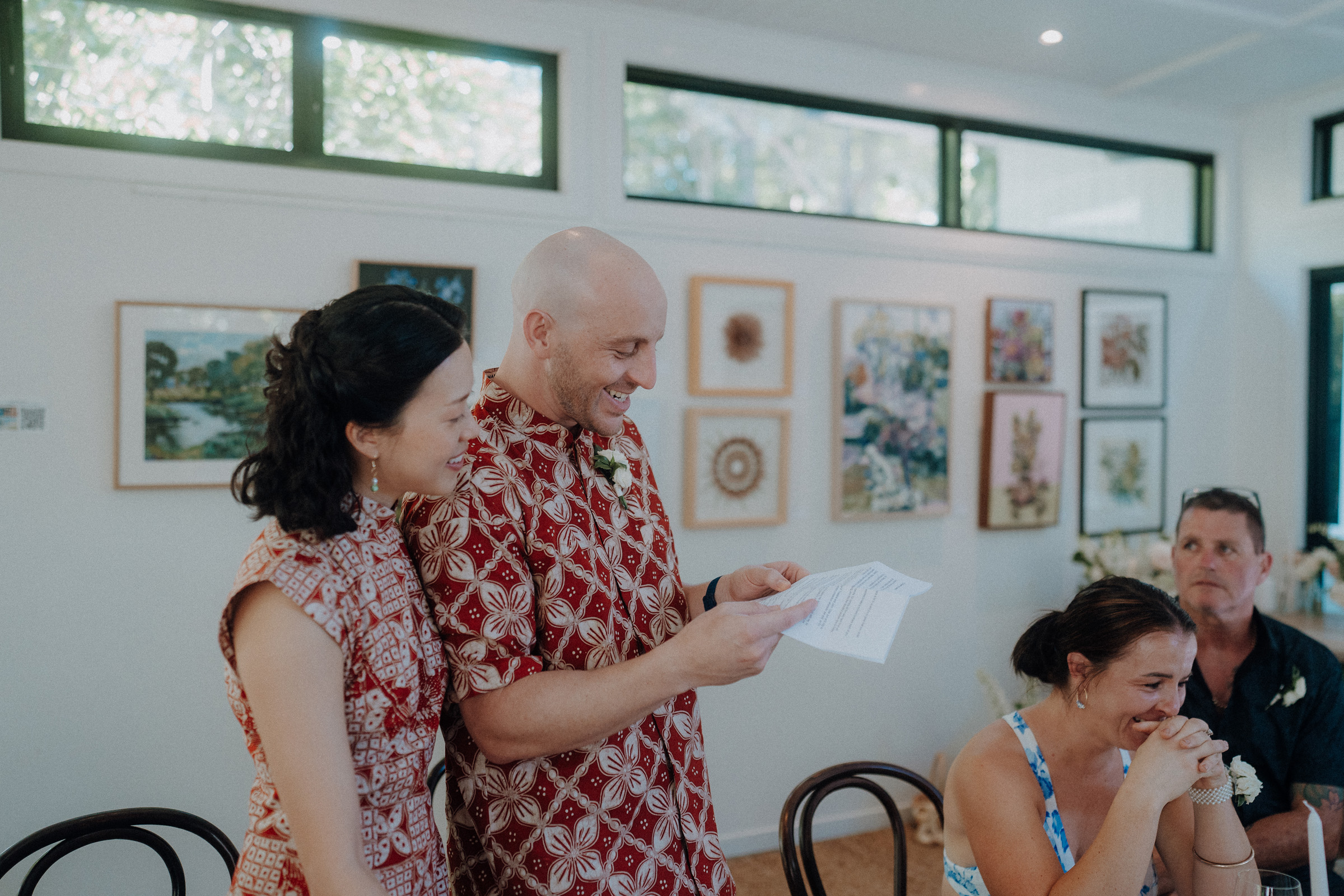 A man and woman in patterned clothing stand and read from paper in an art-filled room. Two seated people listen, one smiling.