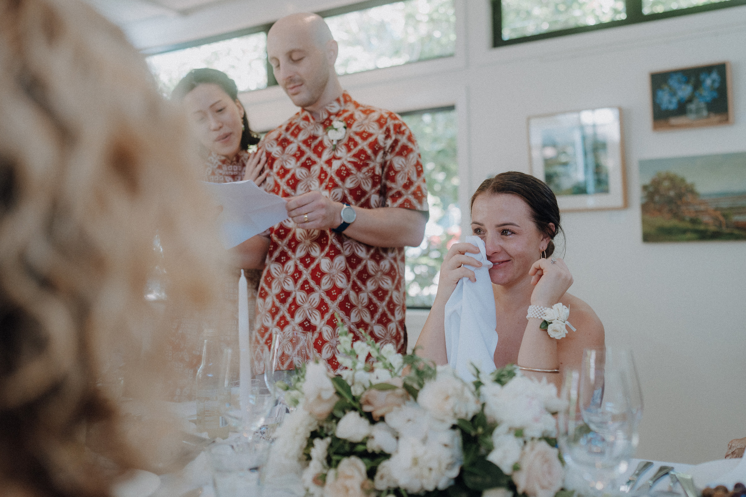 A woman sits at a table with flowers, wiping tears with a tissue. In the background, a man and another woman read from a paper.