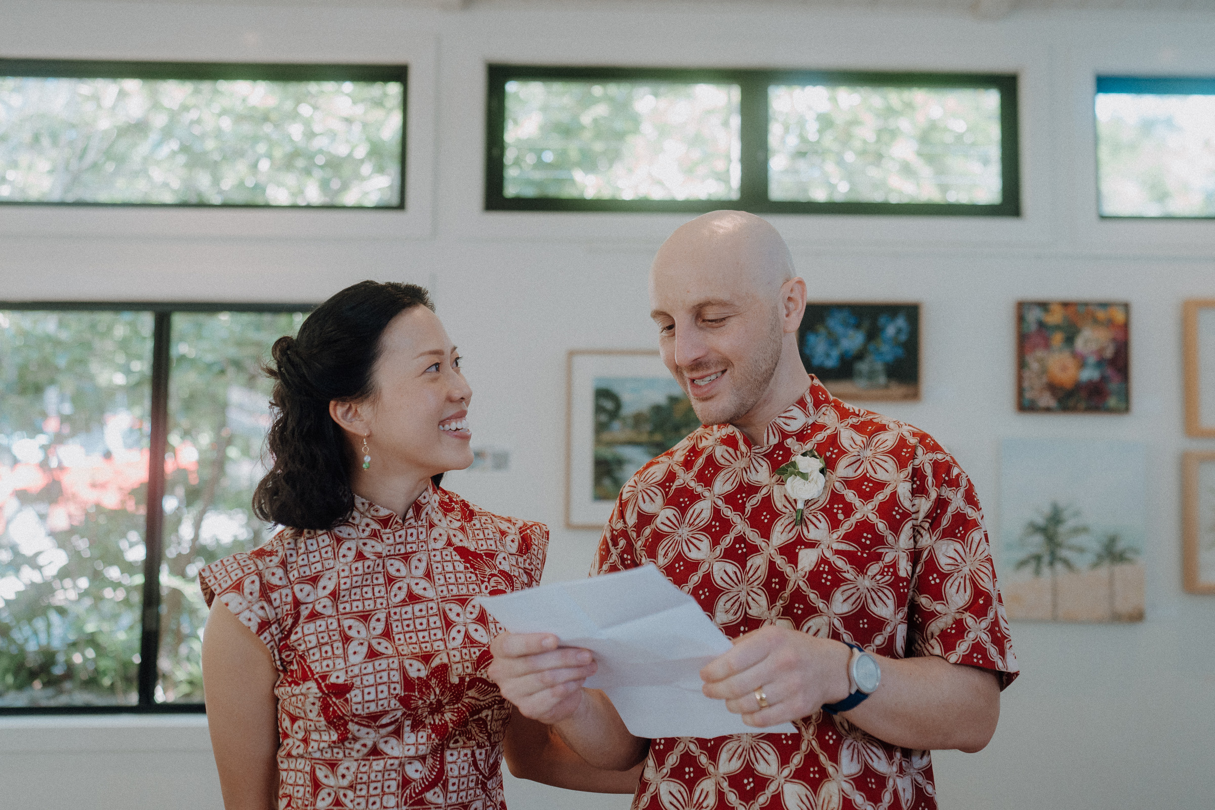 A couple in matching patterned outfits stands indoors; the man holds a piece of paper while they both smile.