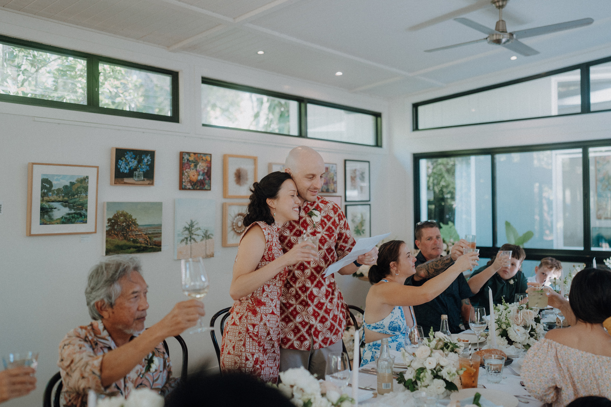 A couple stands and raises a toast during a gathering in a bright room, surrounded by seated guests and floral table arrangements.