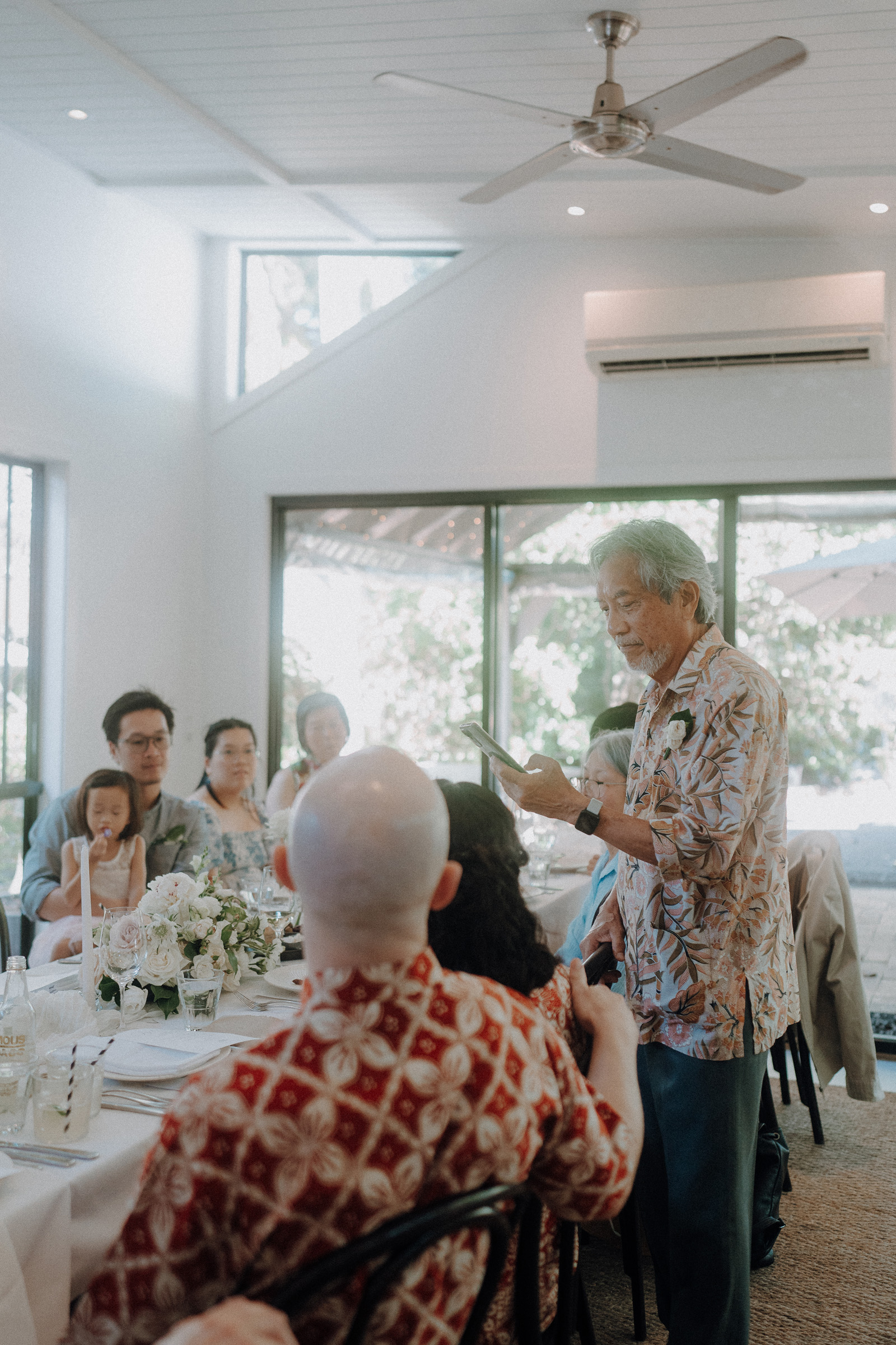 An older man in a floral shirt speaks to seated guests at a long dining table in a bright room with large windows.