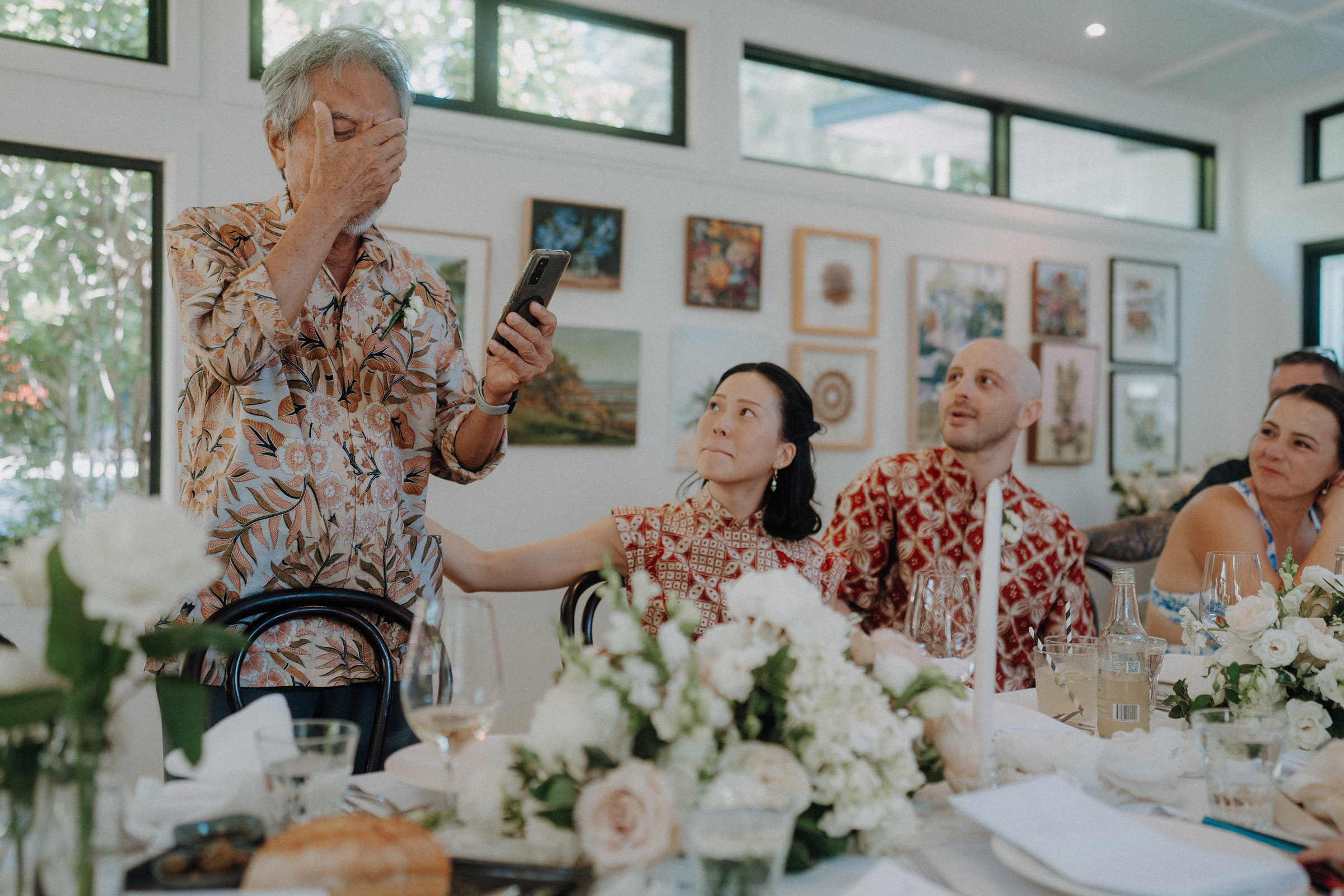 A man stands and wipes his eyes while holding a phone during a speech at a gathering. Three others sit nearby, with one woman looking up at him. The room has floral decor and framed art.