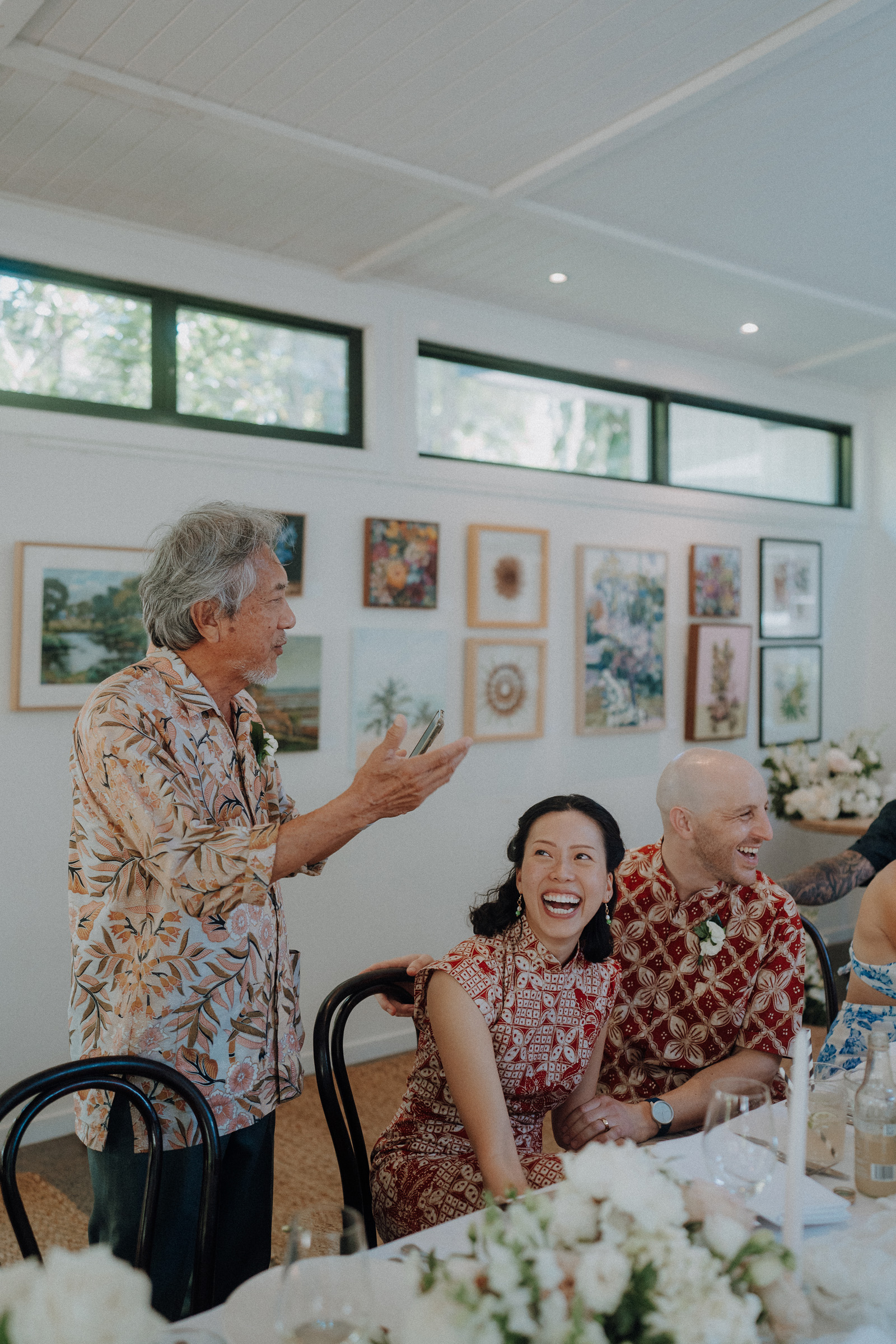 An older man speaks standing next to a seated woman and man, all smiling. They wear patterned clothing in a bright, decorated room with framed pictures on the wall.