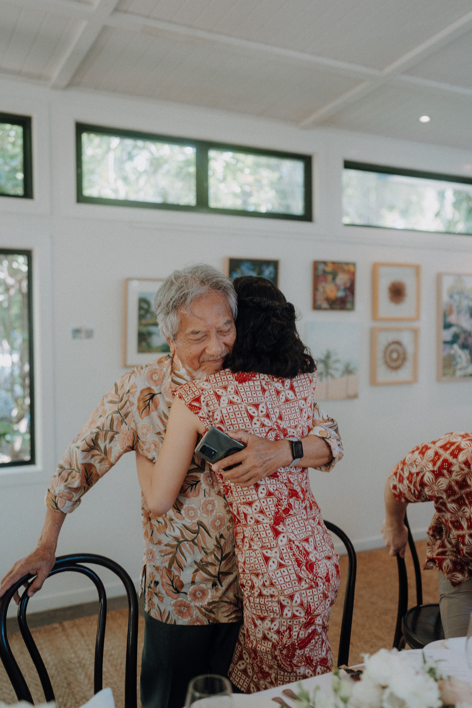 An older man and a woman hug in a bright room with framed pictures on the wall. She holds a phone in one hand.