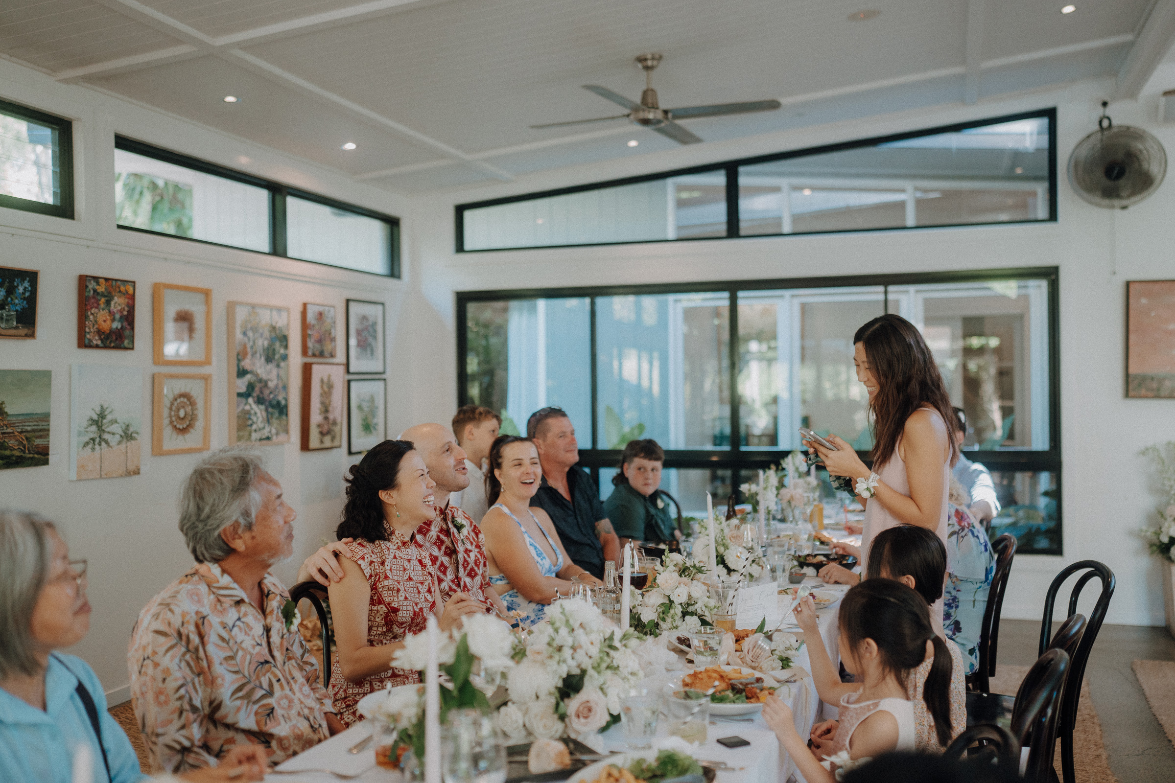 A woman stands speaking to a seated group of people around a long dining table adorned with flowers in a well-lit room.