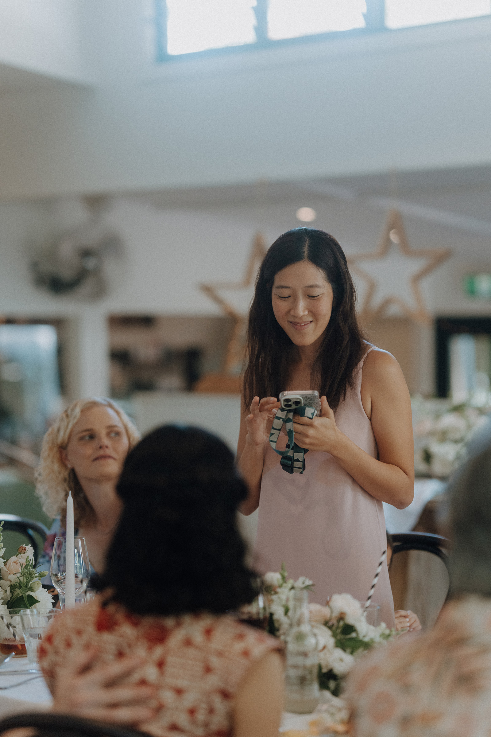 A woman stands and smiles while looking at her phone during a gathering. She is surrounded by seated people and floral decorations.