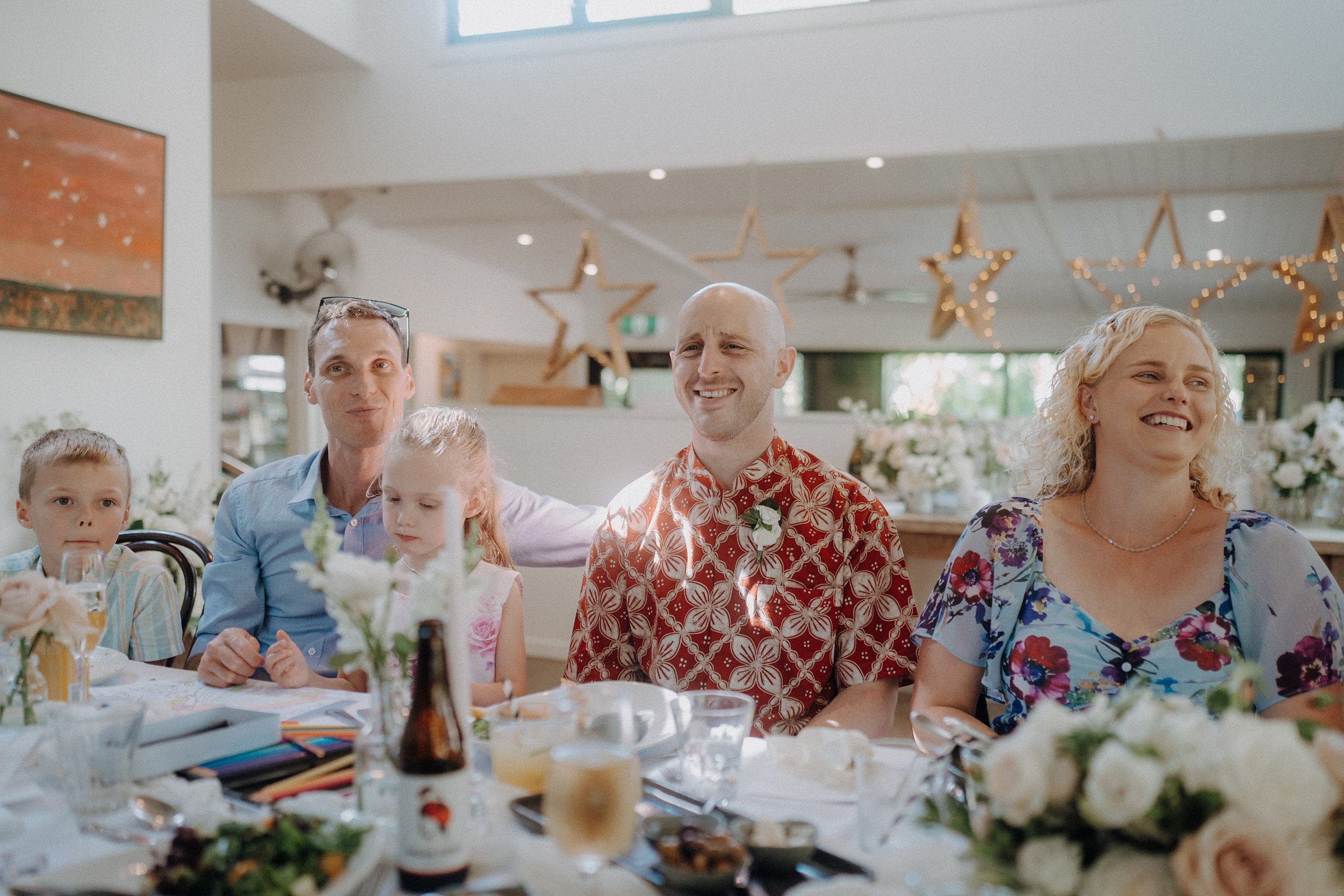 A group of people, including two children, are sitting at a decorated table with food and drinks. They appear to be celebrating, with floral arrangements and star decorations in the background.