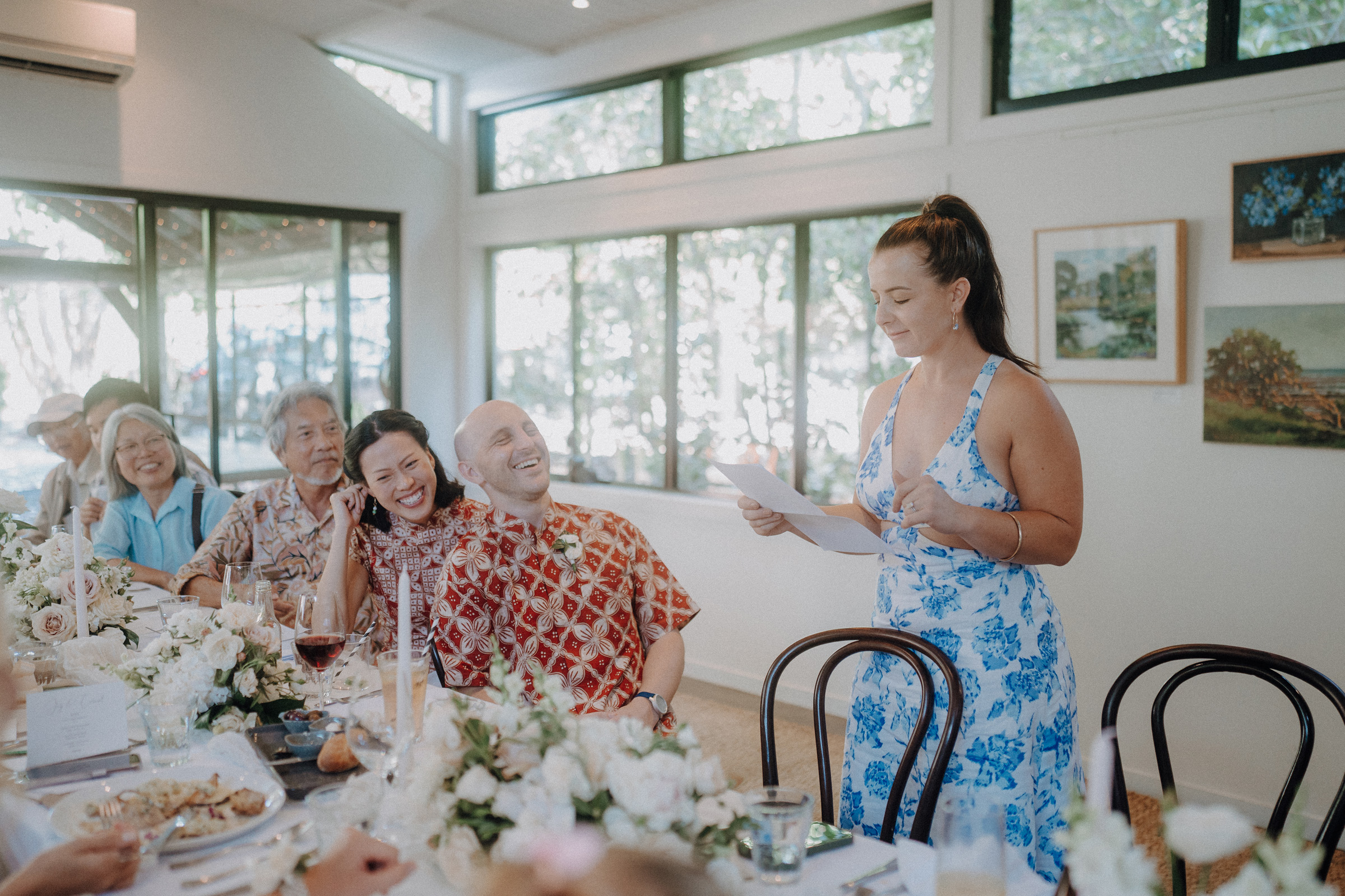 A woman in a blue dress speaks to a seated group at a dining table decorated with white flowers. Others listen and smile in a room with large windows.